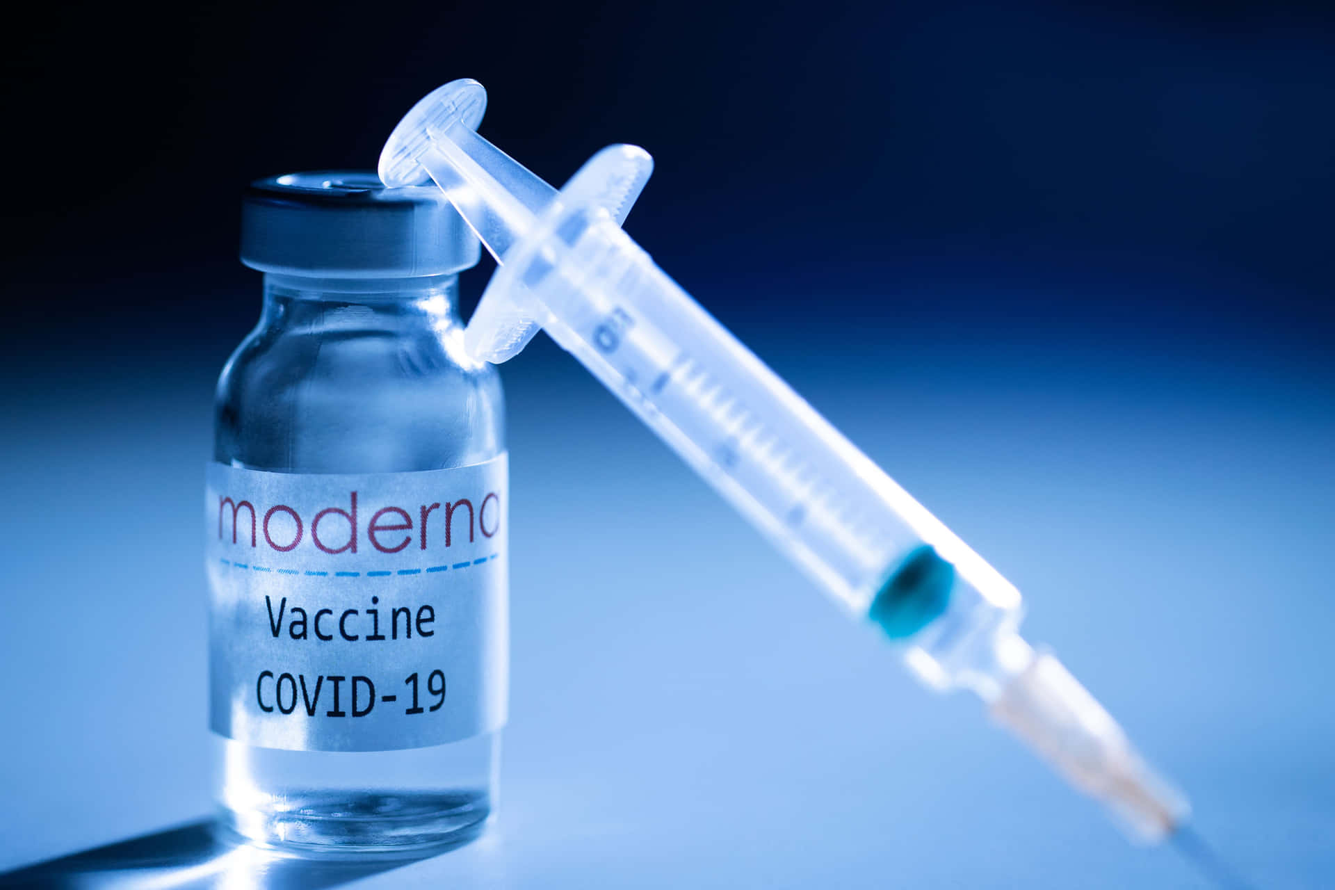 在藍色燈光下，一支注射器靠在 Moderna COVID-19 疫苗瓶上。