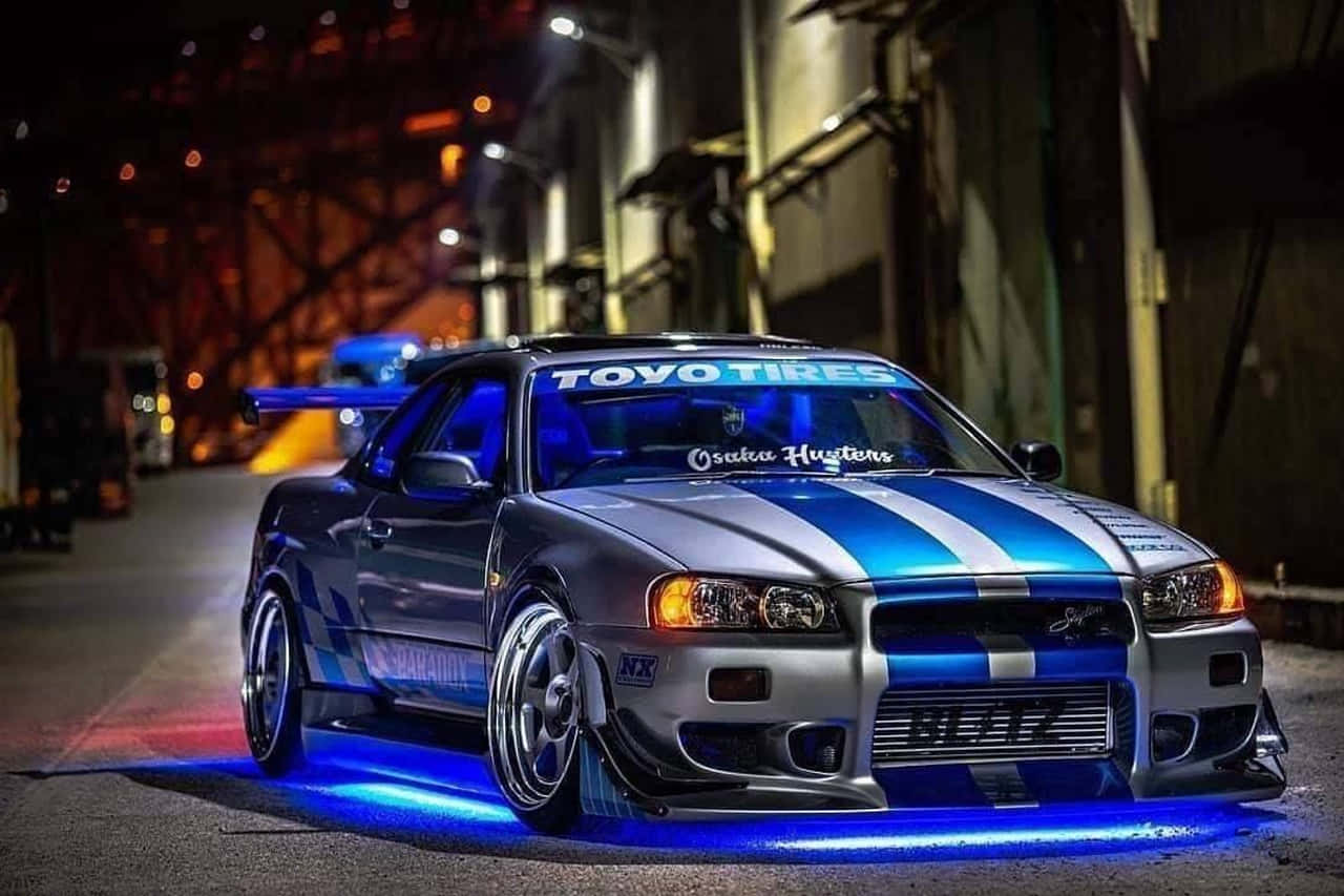 保羅·沃克駕駛Nissan Skyline 壁紙 — 一架改裝的日產Skyline GTR，帶有藍色和銀色條紋，在夜間於粗糙的城市小巷中霓虹燈下發光。