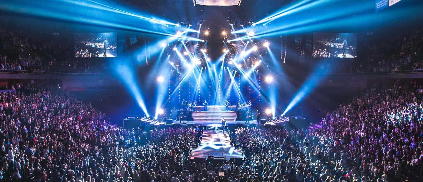 Mohegan Sun Arena 音樂會照片 壁紙 — 在擠滿人的場館中，大量演唱會人群在耀眼的藍色舞台燈光和光束下湧動。