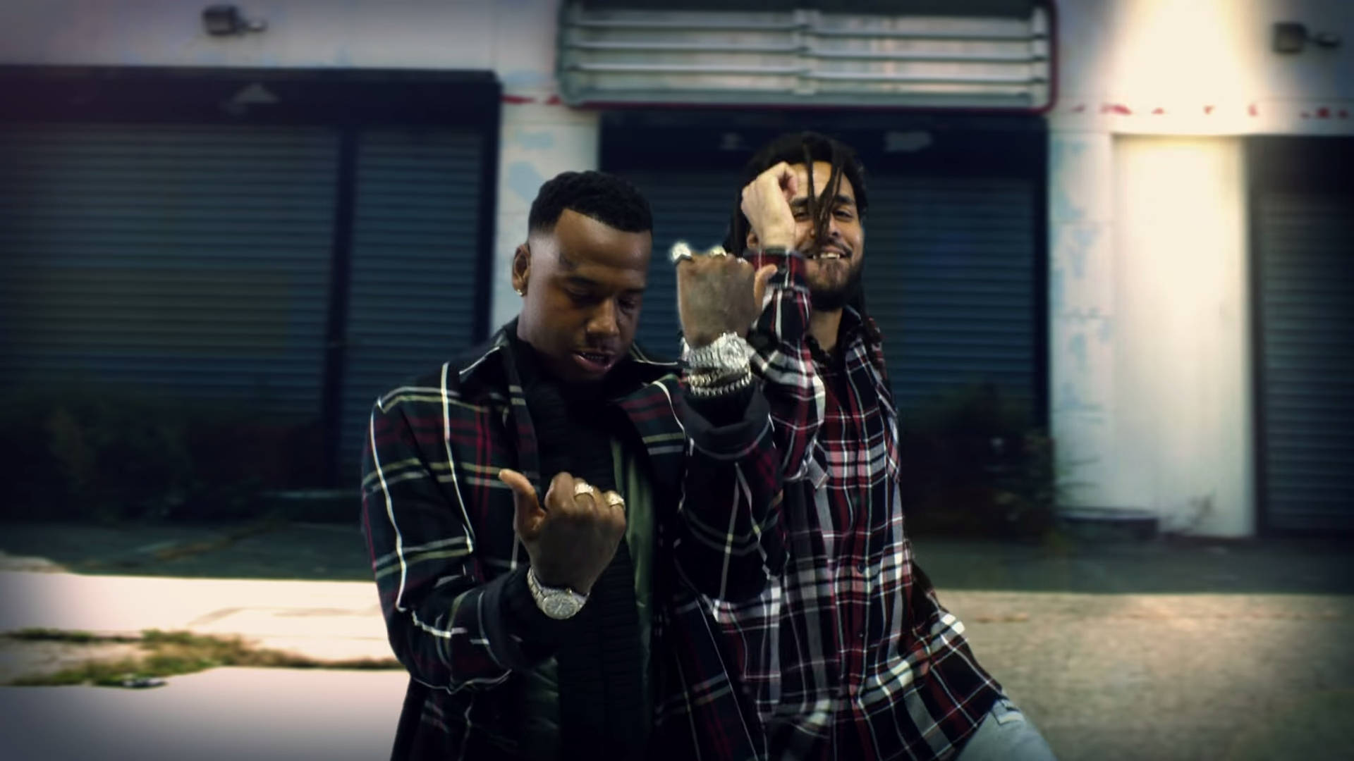 Moneybagg Yo 與 J Cole 壁紙 — 兩個穿著格仔衫嘅男人喺夜晚喺關閉嘅車庫前自信地擺出姿勢，配合手勢。