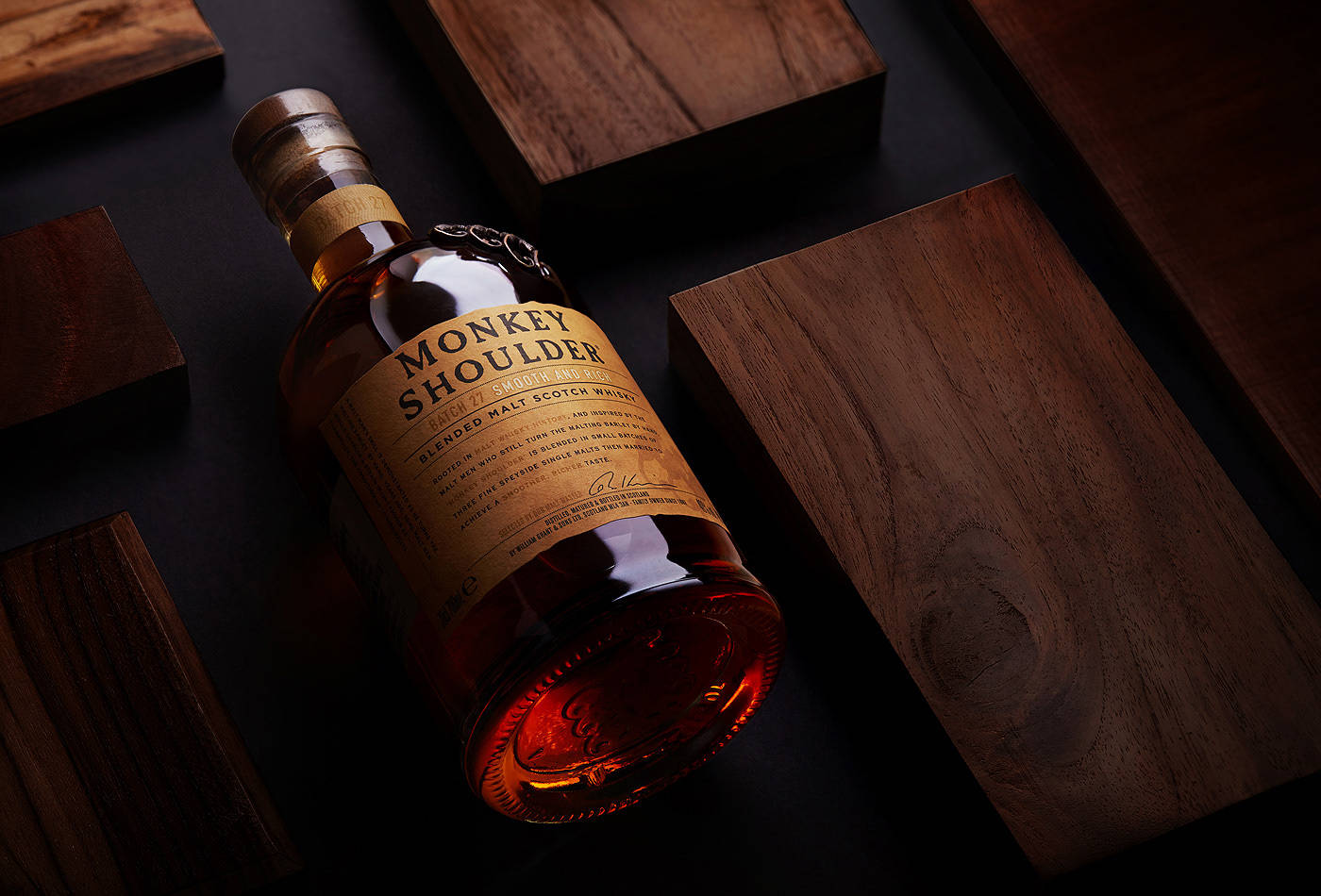 Monkey Shoulder Batch 27，三種單一麥芽威士忌的精緻融合 壁紙 — 一瓶Monkey Shoulder威士忌在戲劇性燈光下斜放在深色木塊上。