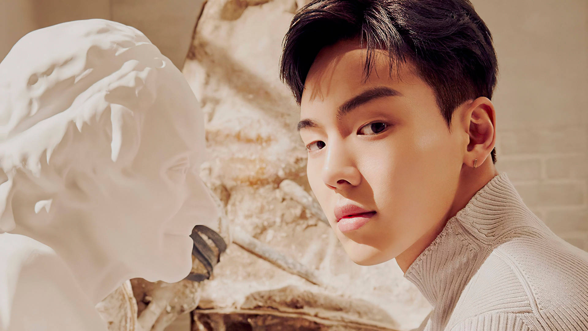 Monsta X Shownu 石像 壁紙 — 一位身穿米色高領毛衣的年輕男子，在溫暖的金色光線下，若有所思地站在古典大理石半身像旁。