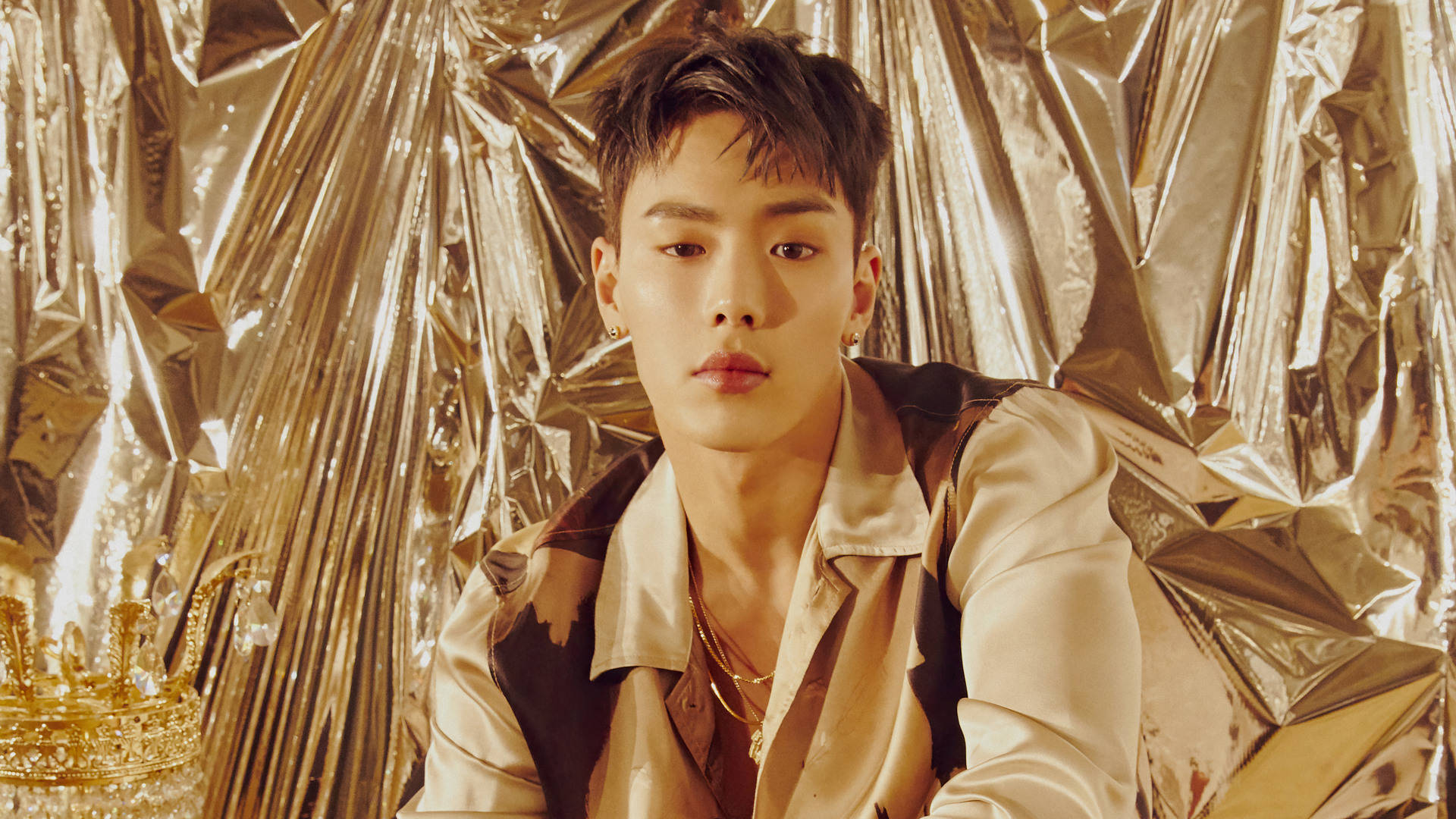 Monsta X Shownu 金屬風 壁紙 — 一位年輕男子，頭髮造型時尚，佩戴金色珠寶，自信地坐在皺褶的金箔背景前，旁邊放著一頂皇冠。