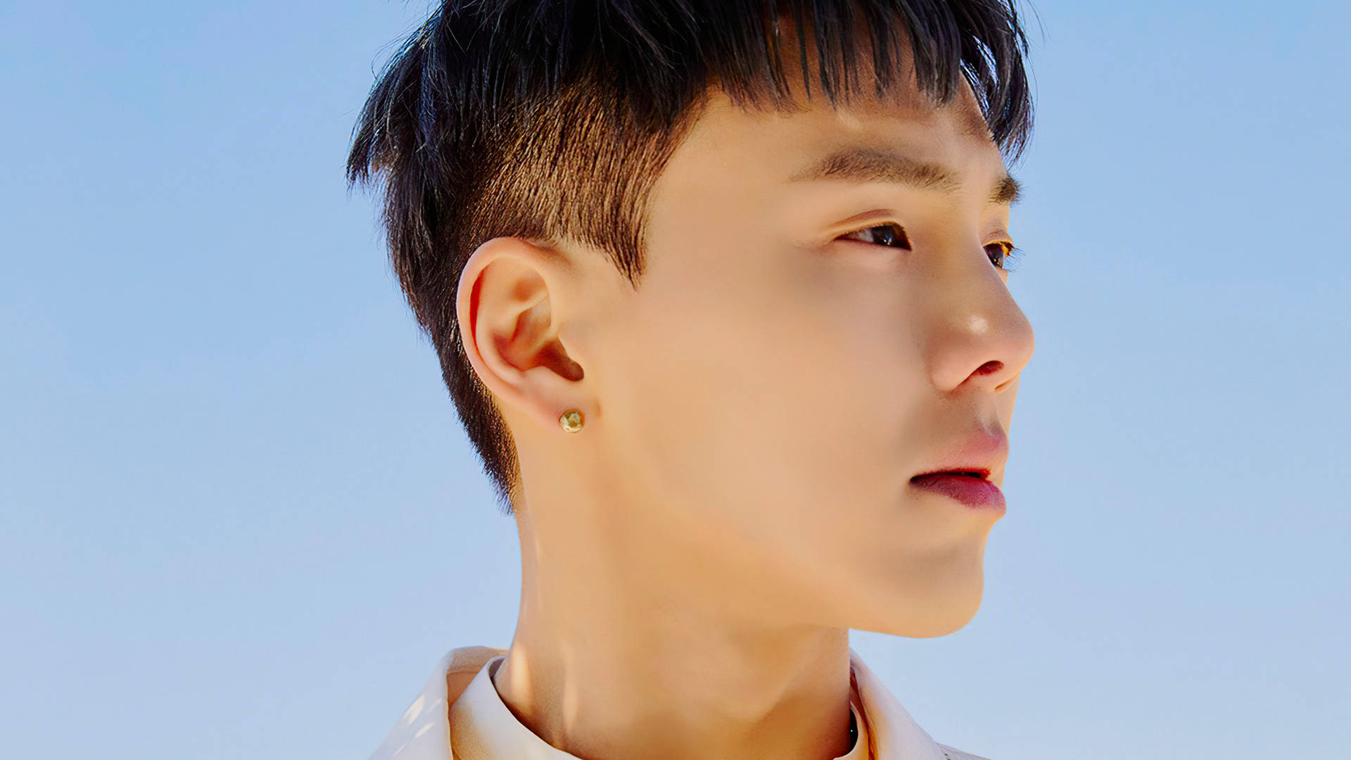 Monsta X Shownu Fantasia X 壁紙 — 一位短黑髮、戴金耳環的年輕男子，在清澈的藍天下若有所思地凝視著。