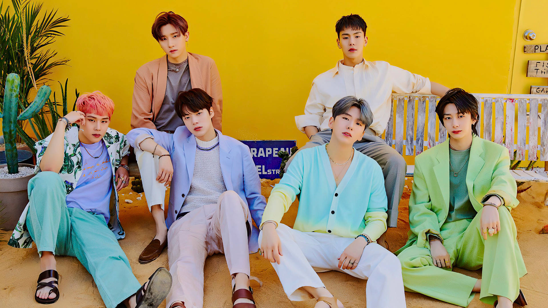 Monsta X 熱帶概念 壁紙 — 六位時尚年輕男子身穿色彩繽紛的服裝，在明亮的黃色牆壁和沙漠植物前自信地擺姿勢。