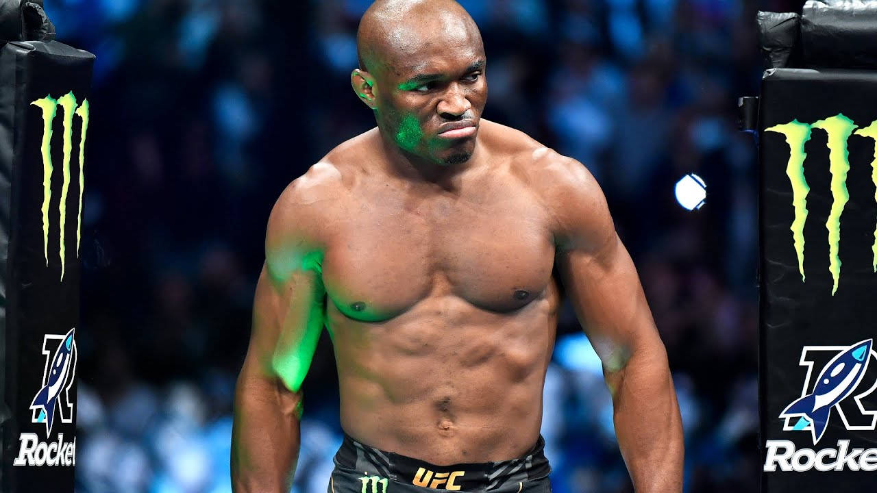 卡馬魯·烏斯曼與Monster Energy 壁紙 — 一位肌肉發達的UFC拳手站在八角籠內，綠色燈光下，背景有模糊的人群和Monster Energy品牌標誌。