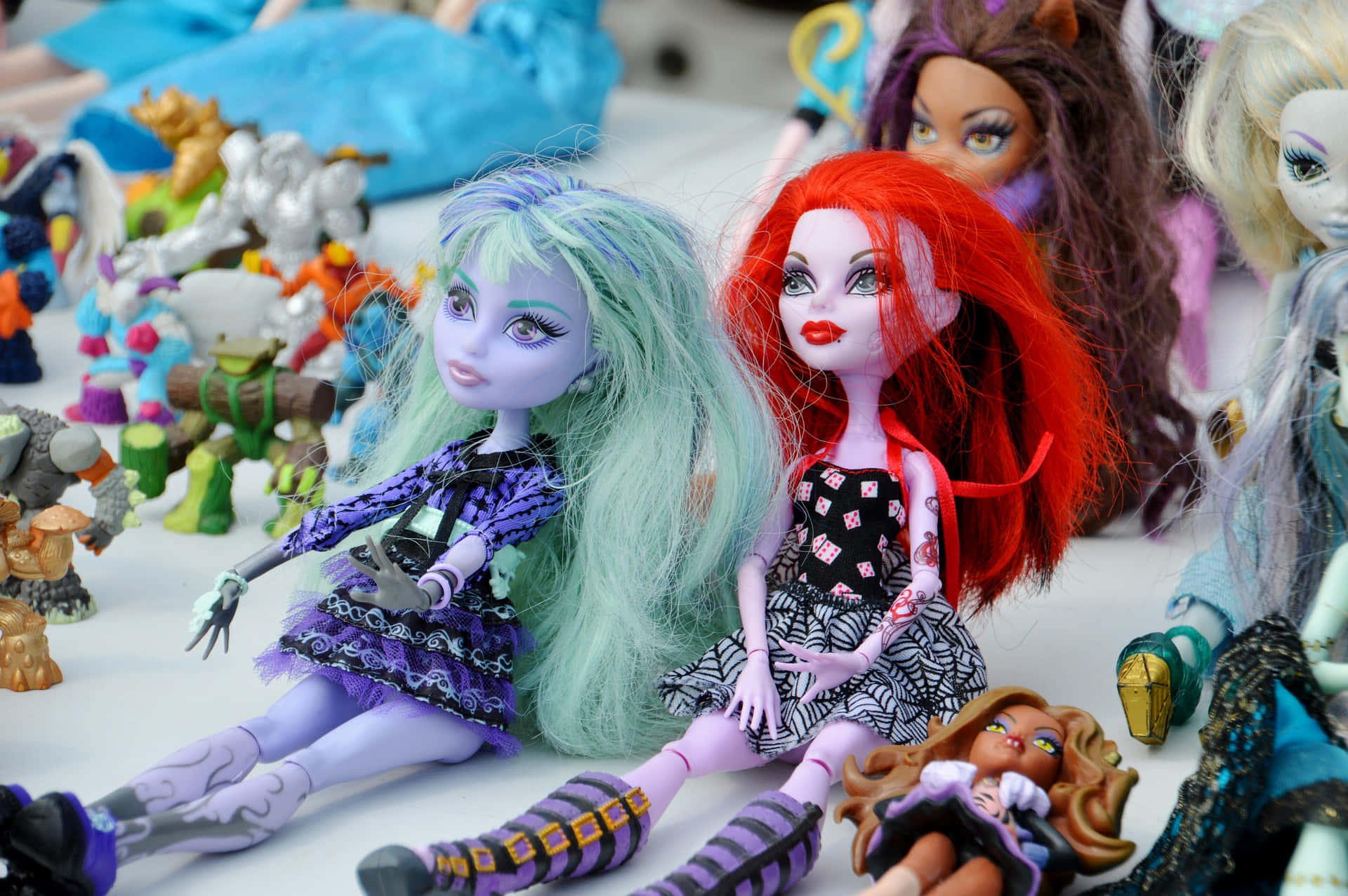 《Monster High》玩偶庭院拍賣場景 壁紙 — 色彩繽紛的Monster High公仔，擁有彩色頭髮和哥德式服裝，坐在白色表面上的各種動作公仔之間。