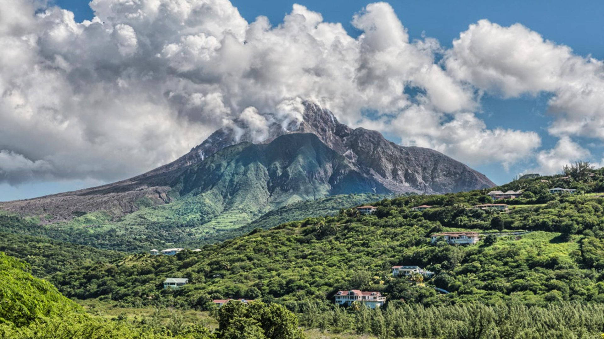 火山在藍天白雲下，於綠色山丘上冒煙，山丘上散落著房屋。
