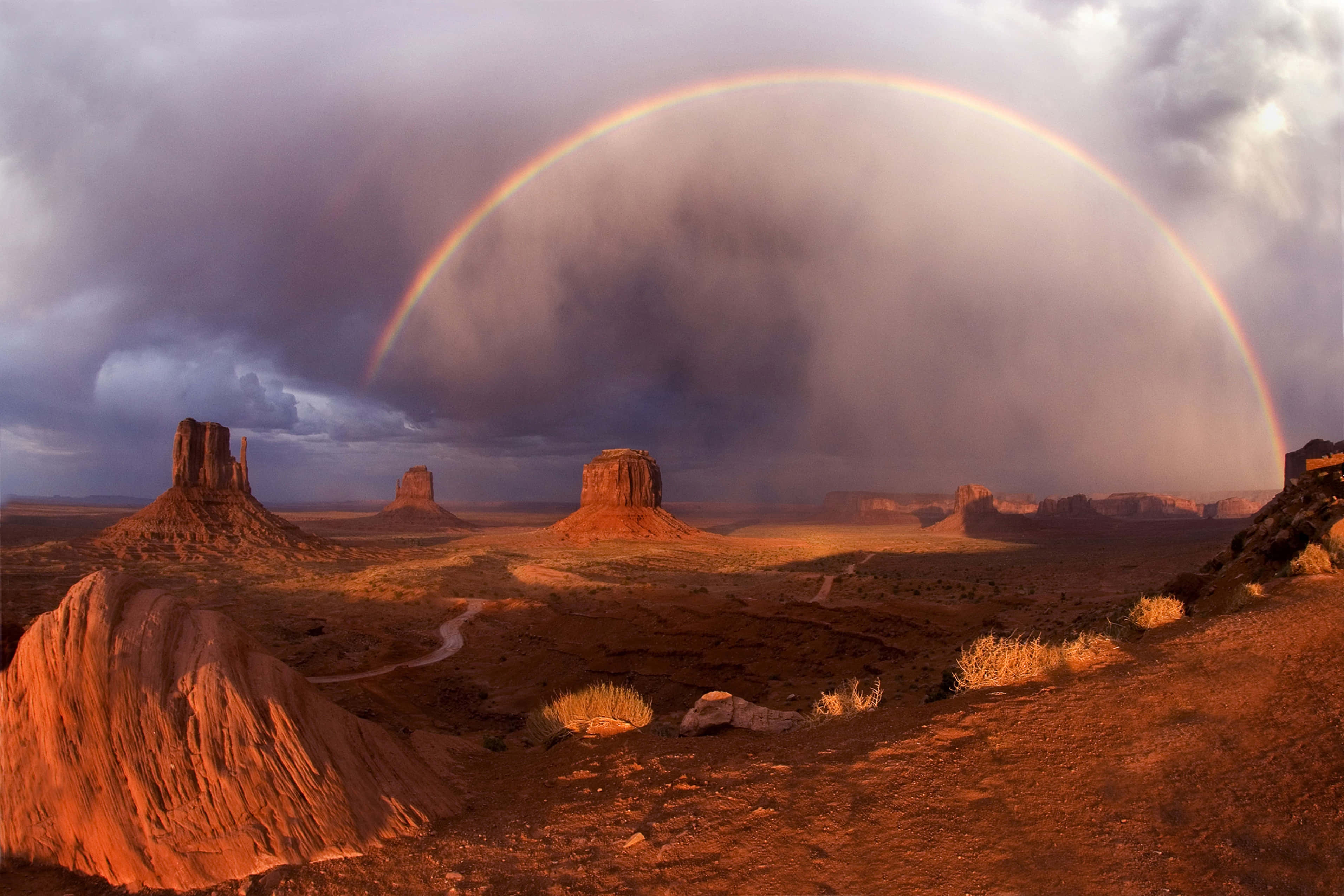 Monument Valley納瓦霍部落公園彩虹 壁紙 — 在暴風雨的天空下，一道鮮豔的彩虹在沙漠景觀中戲劇性的紅色岩層上拱起。
