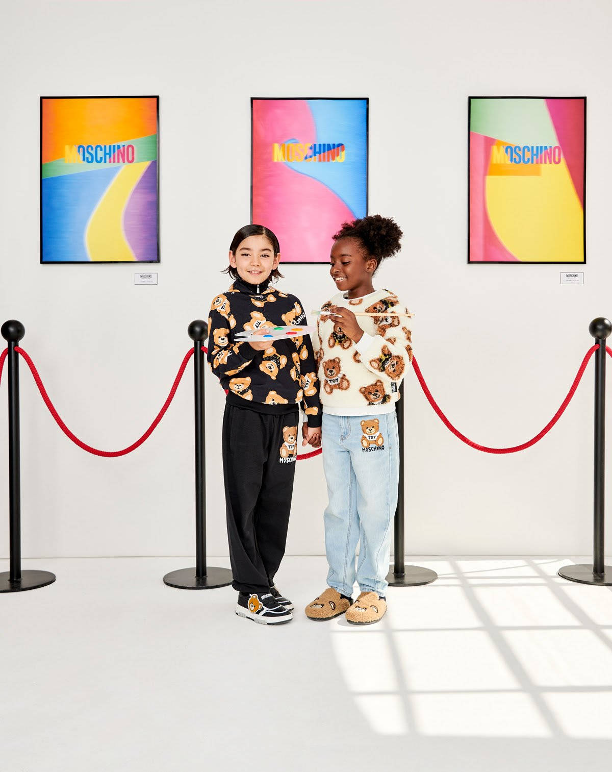 Moschino童趣畫廊 壁紙 — 兩個穿著 Moschino 泰迪熊毛衣的孩子在畫廊裡微笑，畫廊裡有色彩繽紛的抽象藝術和紅色天鵝絨繩索。