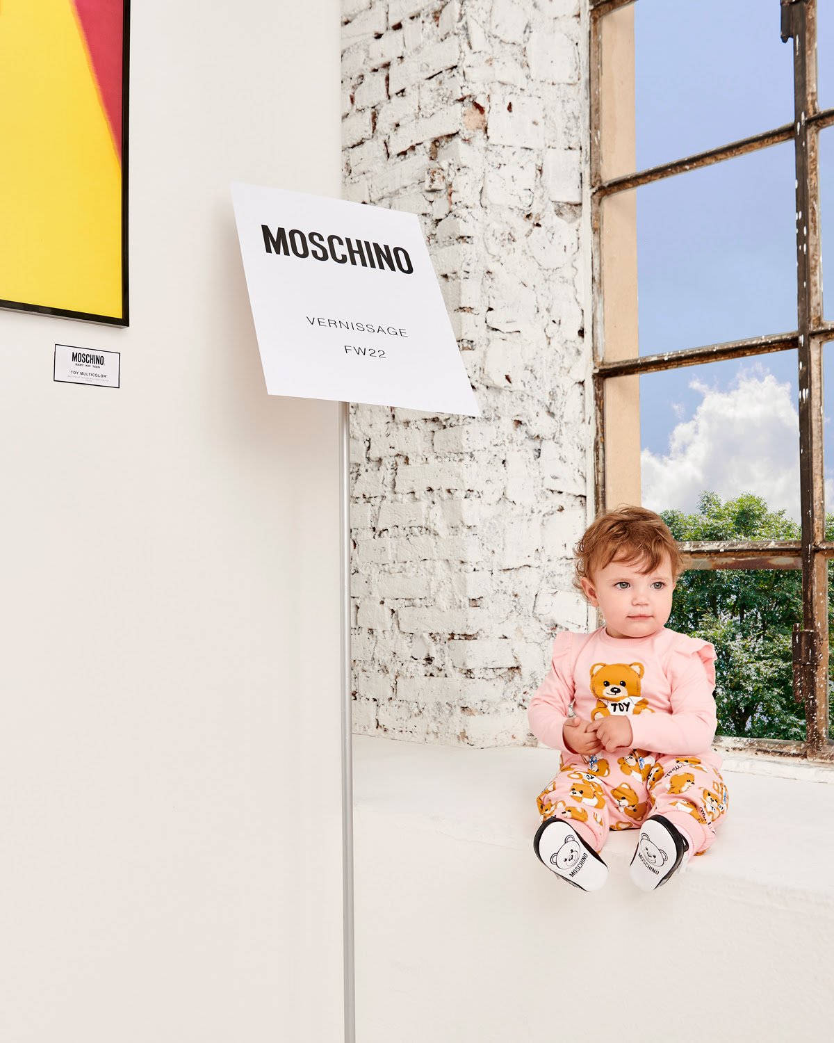Moschino 嬰兒窗邊 壁紙 — 一個穿著粉紅色小熊睡衣的嬰兒坐在白色窗台上，旁邊是一扇飽經風霜的窗戶，窗外是明亮的藍天。