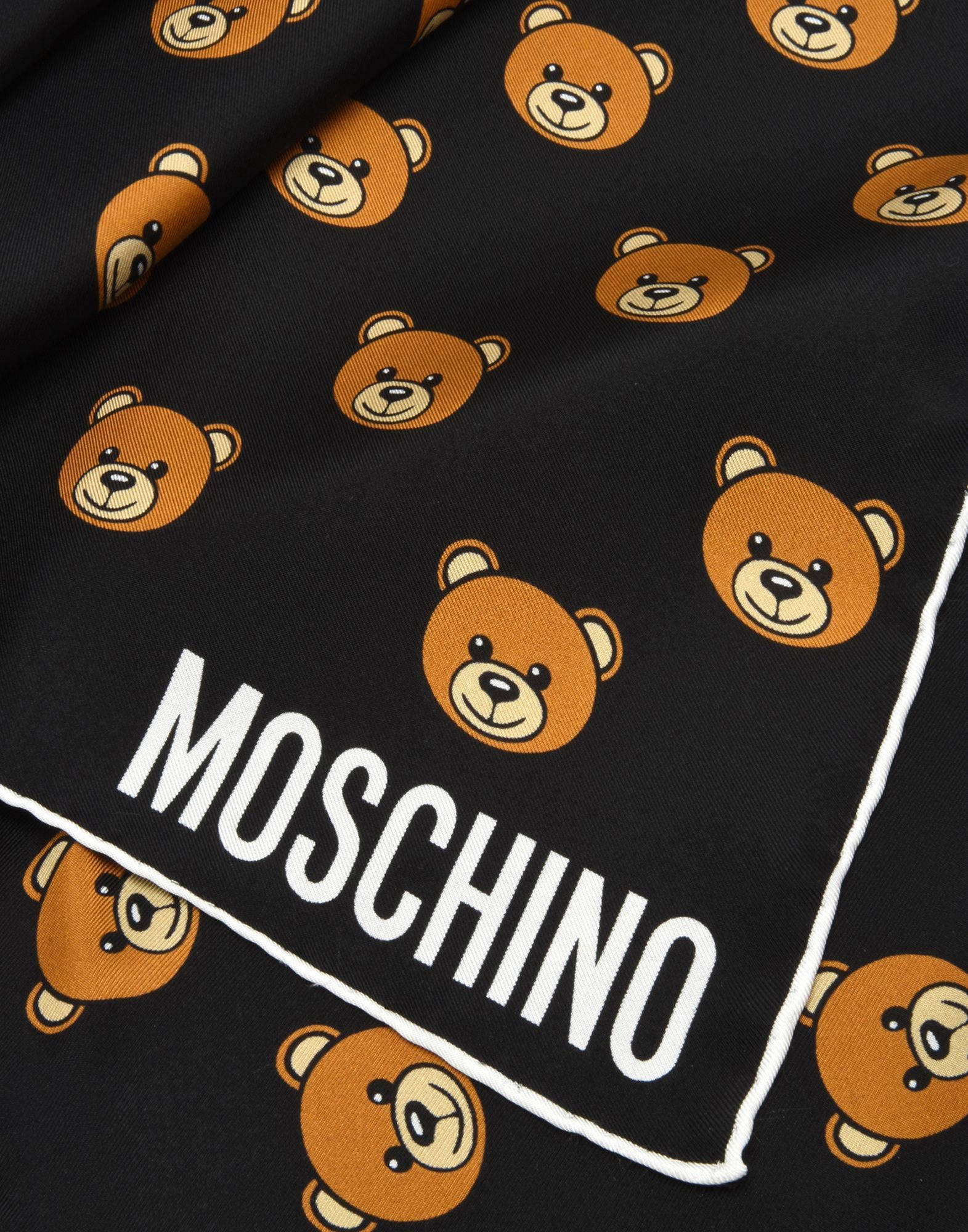 Moschino 泰迪熊圖案布料 壁紙 — 黑色布料圍巾的特寫，帶有重複的橙色泰迪熊臉和粗體白色MOSCHINO品牌標誌。