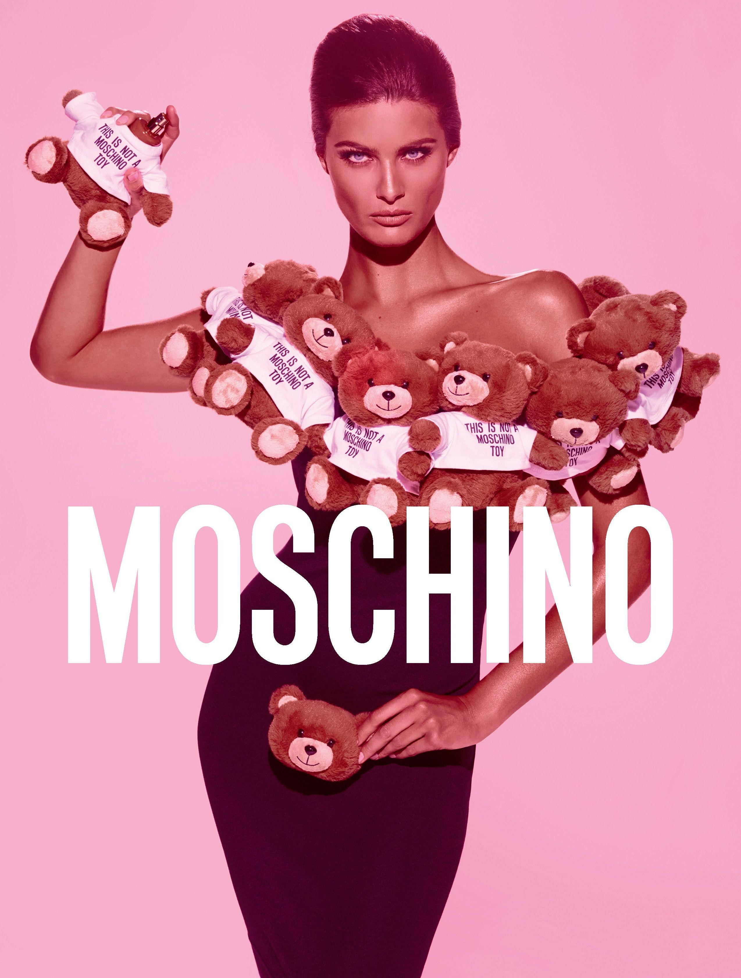 Moschino 泰迪熊連衣裙 壁紙 — 一位穿著黑色連衣裙的模特兒在粉紅色背景前抱著穿著「this is not a moschino toy」襯衫的泰迪熊。
