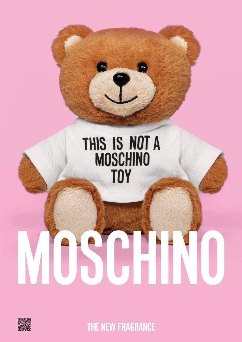 Moschino泰迪熊粉紅背景 壁紙 — 一隻棕色泰迪熊穿著白色T恤，上面有文字，坐在純粉紅色背景前，帶有大型白色品牌標誌。