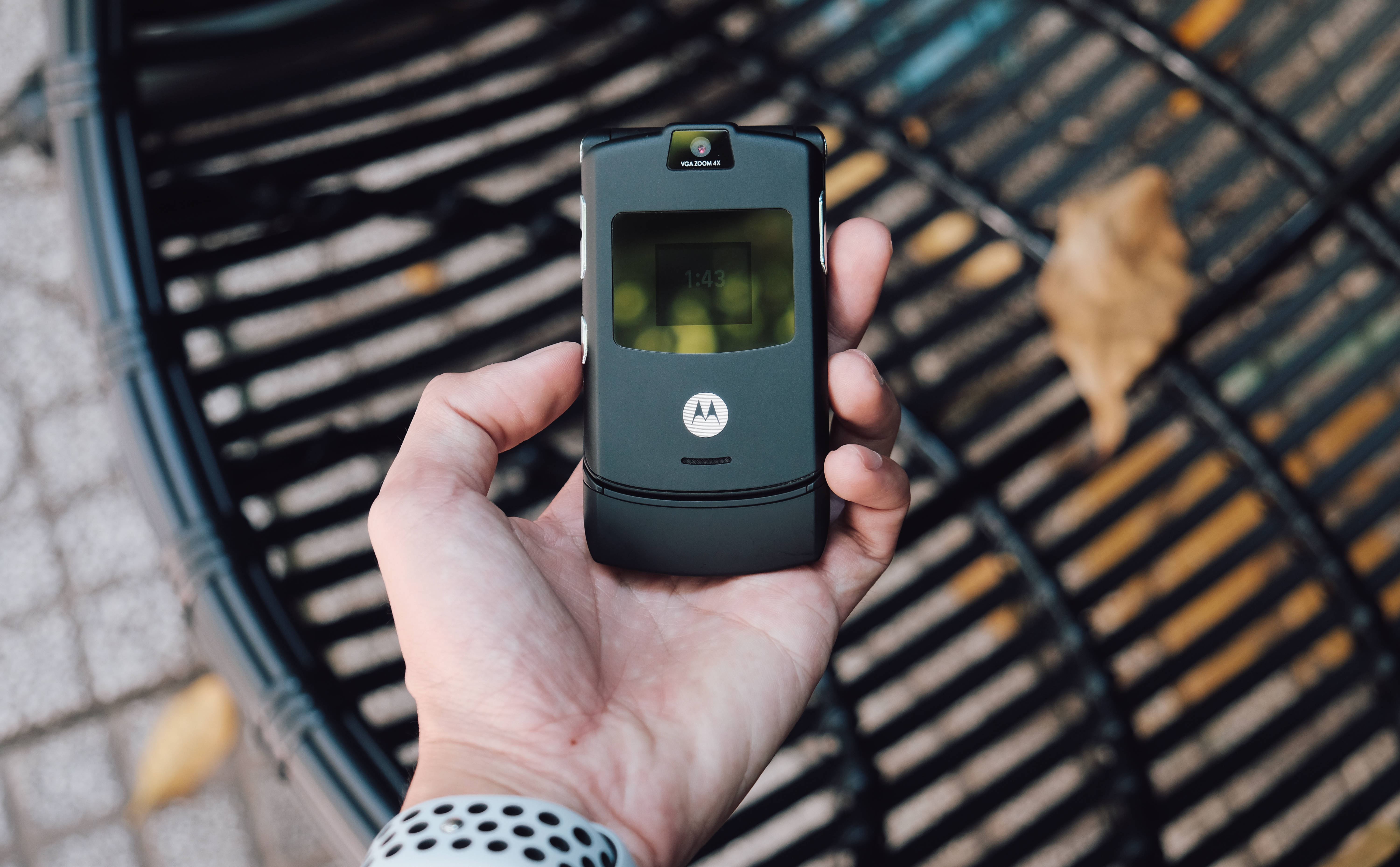 黑色揭蓋 Motorola 壁紙 — 一隻手拿著一部黑色Motorola摺機，屏幕發光，背景是模糊的長椅和落葉。