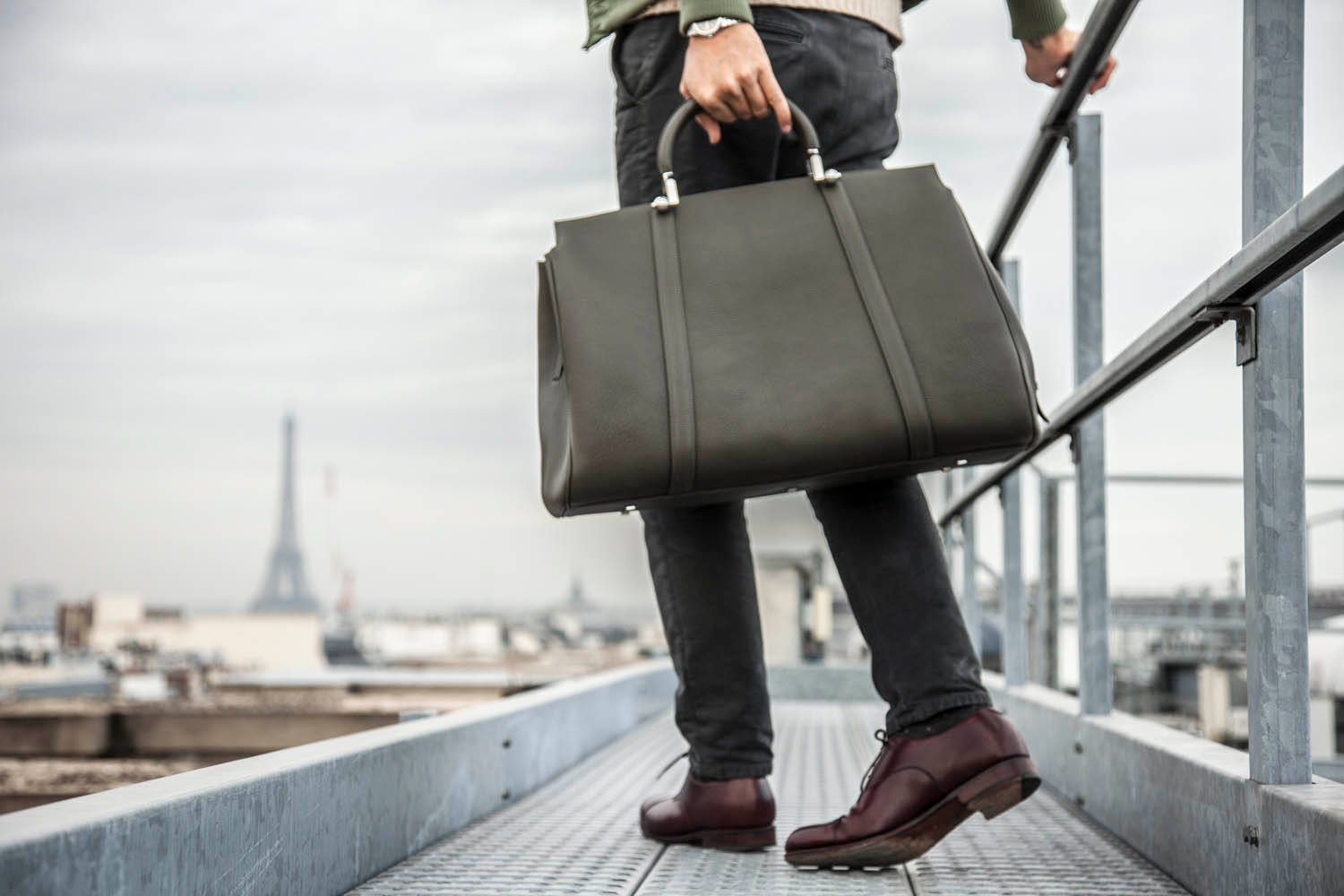 以品質與可靠性打造的 Moynat 男款手袋 壁紙 — 一個人穿著深色褲子和酒紅色鞋子，拿著一個綠色皮革公文包，走在金屬屋頂上，俯瞰巴黎，遠處有艾菲爾鐵塔。