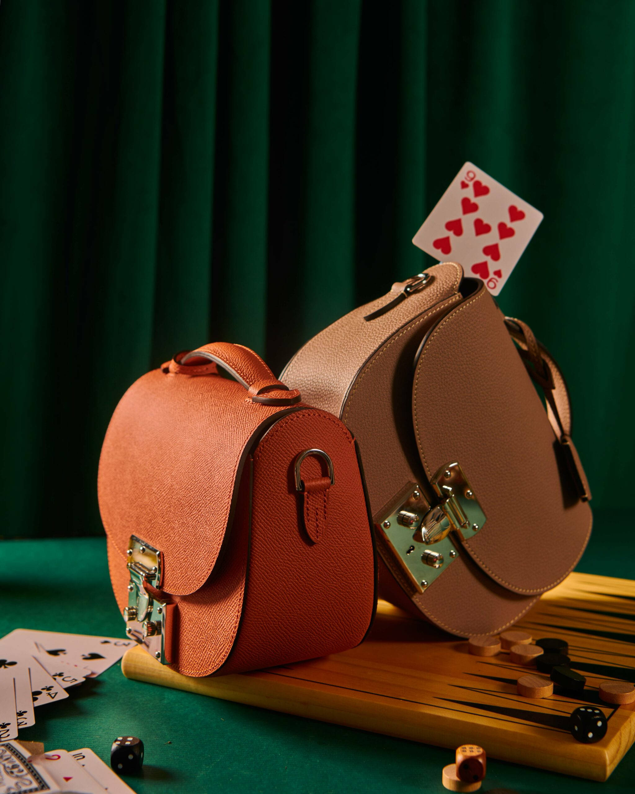 獨家Moynat Flori手袋 壁紙 — 兩個質感皮革公事包放在一個背板上有散落的撲克牌和骰子的雙陸棋盤上，背景是深綠色窗簾。