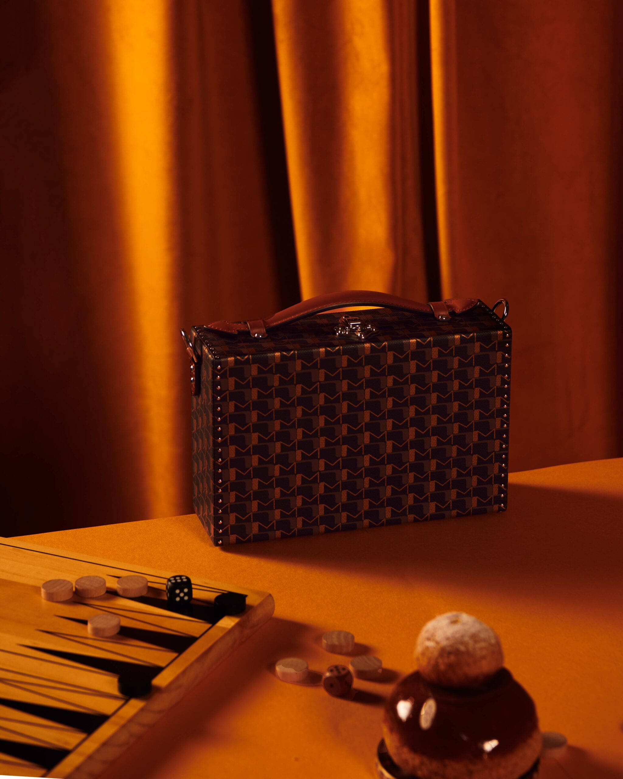 Moynat Monogram公事包 壁紙 — 一個帶圖案的公文包放在桌子上，旁邊有一個雙陸棋套裝和一個朱古力甜品，在溫暖的金色燈光下。