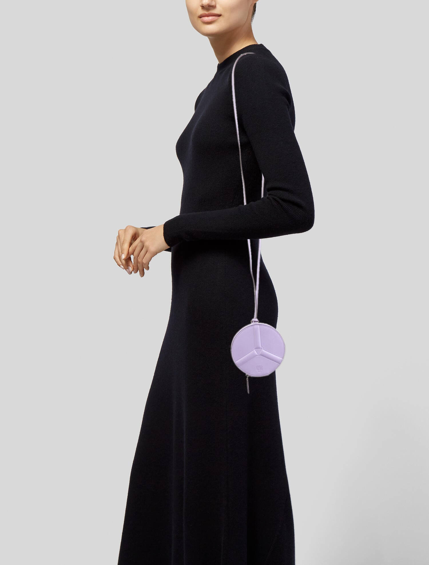 Moynat Zippy Wheel手提包 壁紙 — 一位身穿時尚黑色連衣裙的女性，在灰色中性背景前，手持一個帶有和平標誌設計的淡紫色圓形手袋。