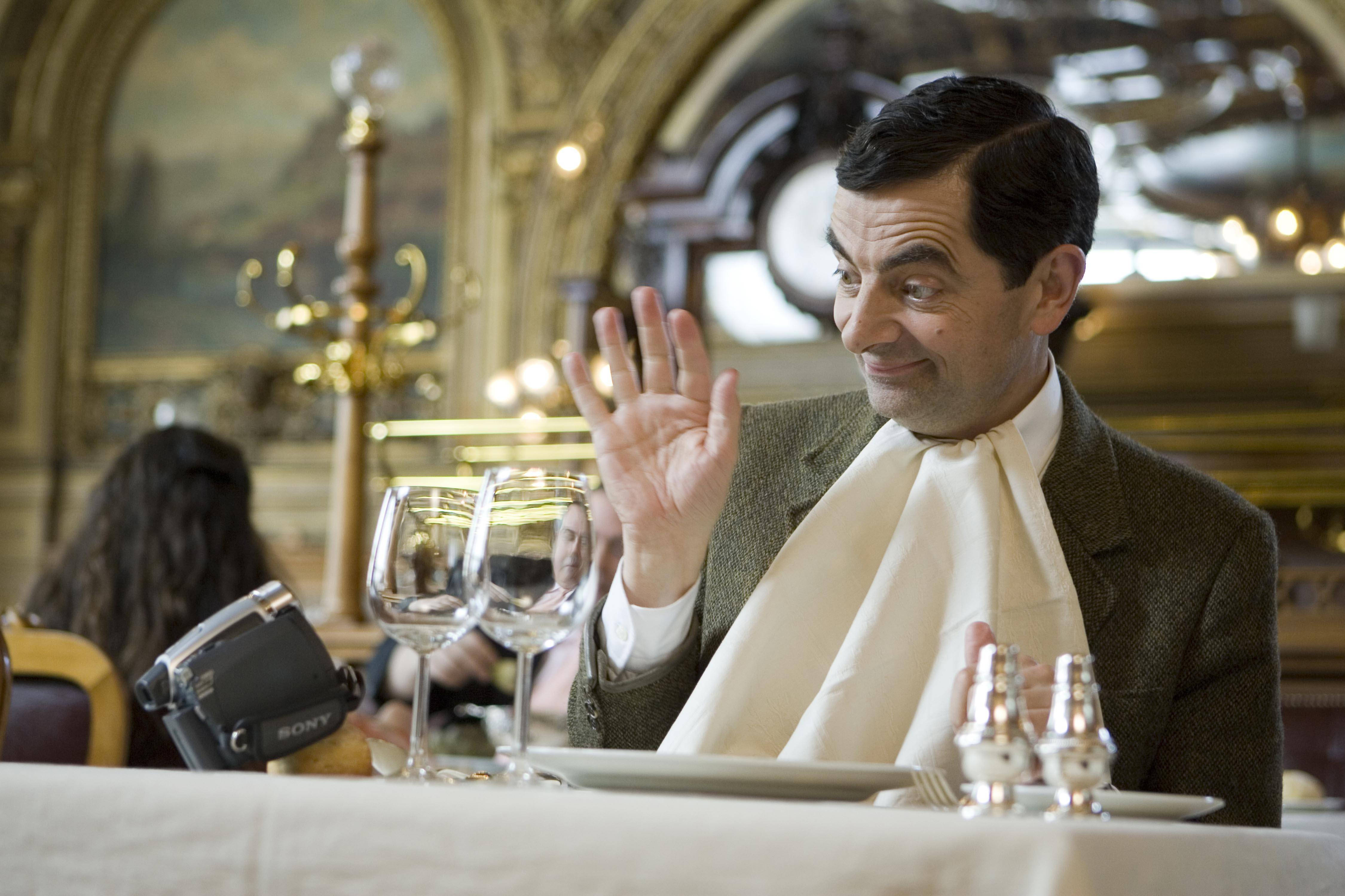 4K 桌布 壁紙 — Mr Bean 喺一個華麗嘅金色裝飾同酒杯嘅高級餐廳枱上開心咁揮手。