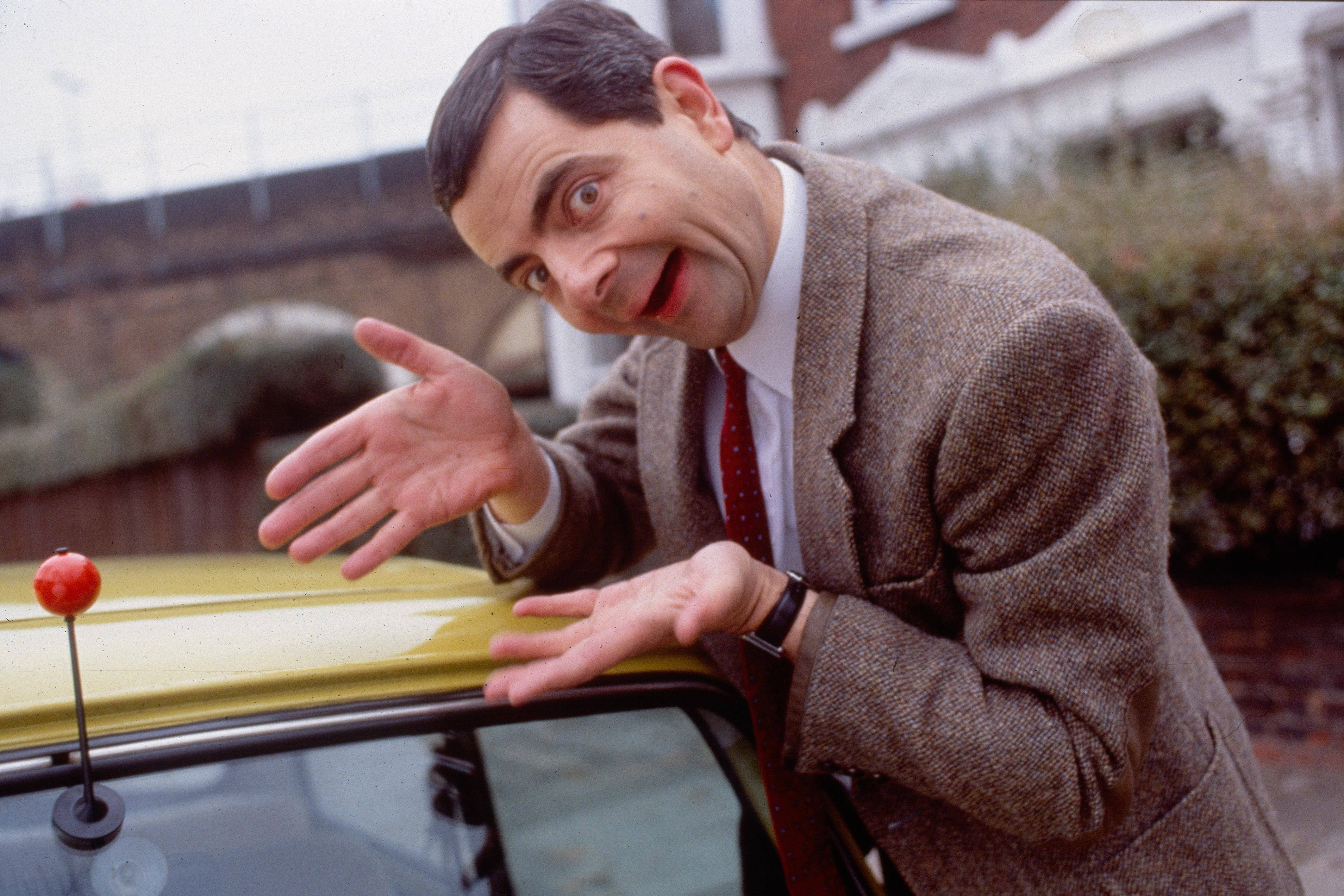 4K 桌布 壁紙 — Mr Bean 喺郊區環境中俯身喺一架黃色車上面，表情滑稽，雙手張開。