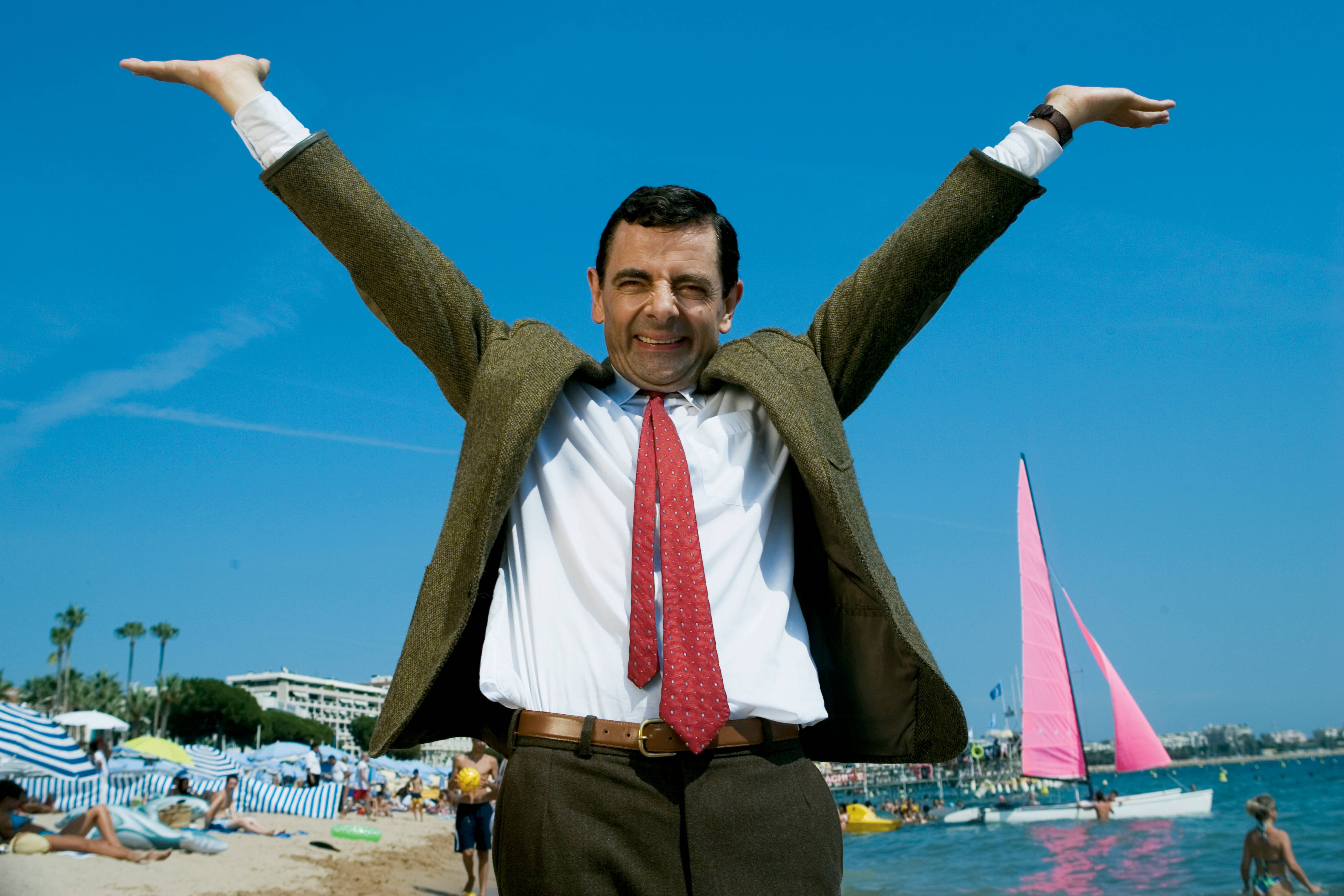 海灘 桌布 壁紙 — Mr Bean 在陽光明媚的海灘上張開雙臂，對著鮮豔的藍天和粉紅色的帆船，開心地站著。