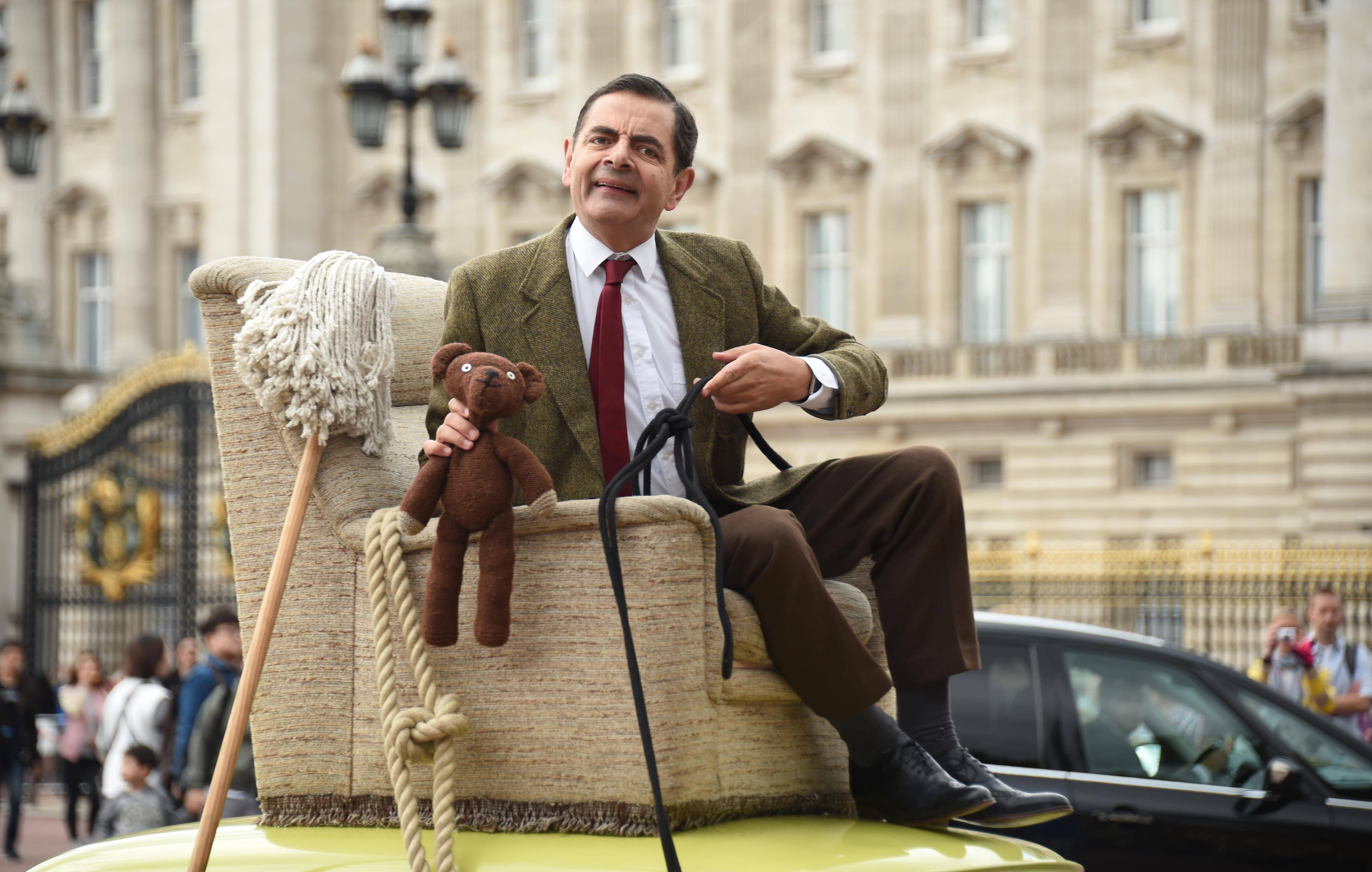 4K 桌布 壁紙 — Mr Bean 坐在一輛黃色車頂上，抱著一隻泰迪熊，旁邊有拖把和繩子，面前是一座宏偉的建築。