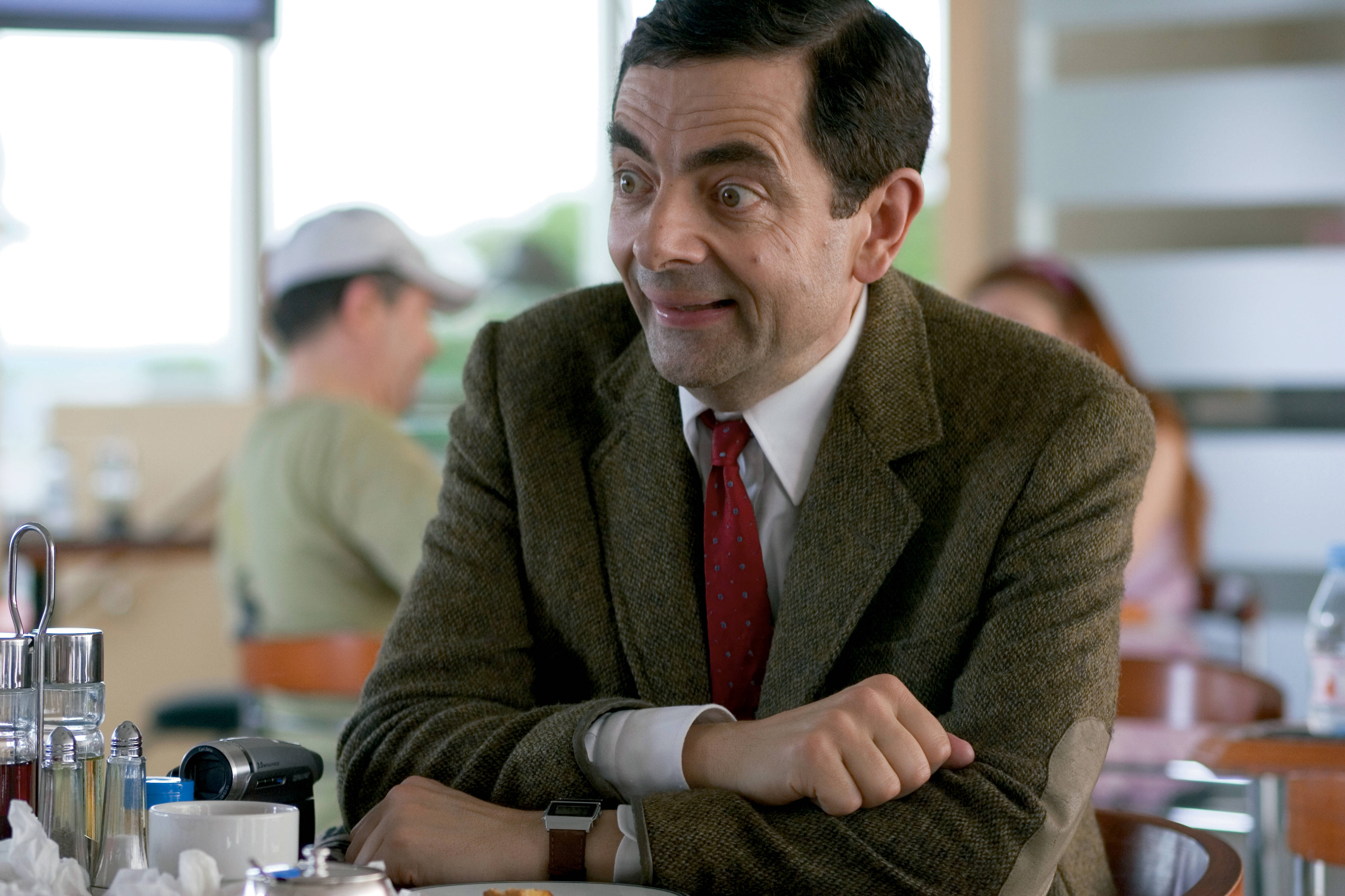 4K 桌布 壁紙 — Mr Bean 坐在咖啡店桌子旁，睜大眼睛，露出頑皮的笑容，穿著棕色粗花呢外套和紅色領帶。
