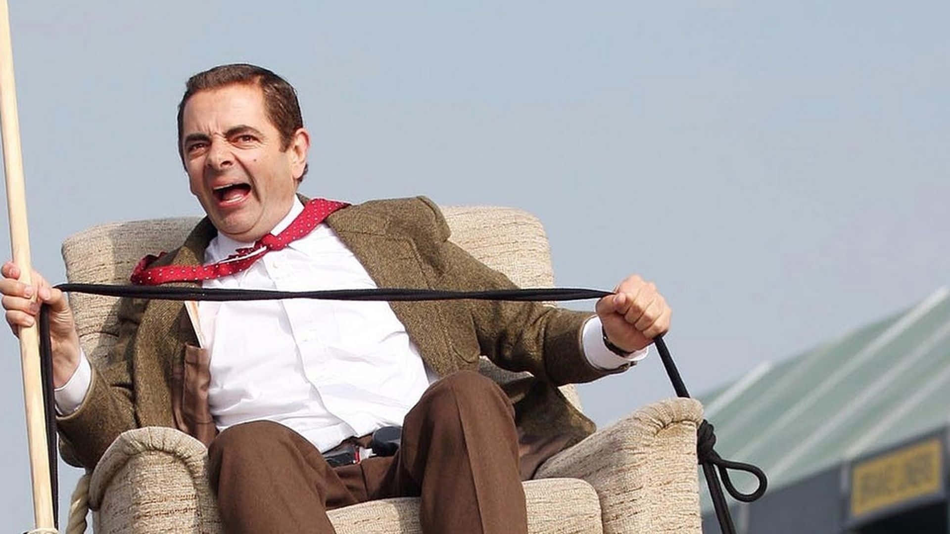 Mr Bean 桌布 壁紙 — Mr Bean 坐在戶外的米色扶手椅上，用力拉著一條黑色繩子，表情滑稽。