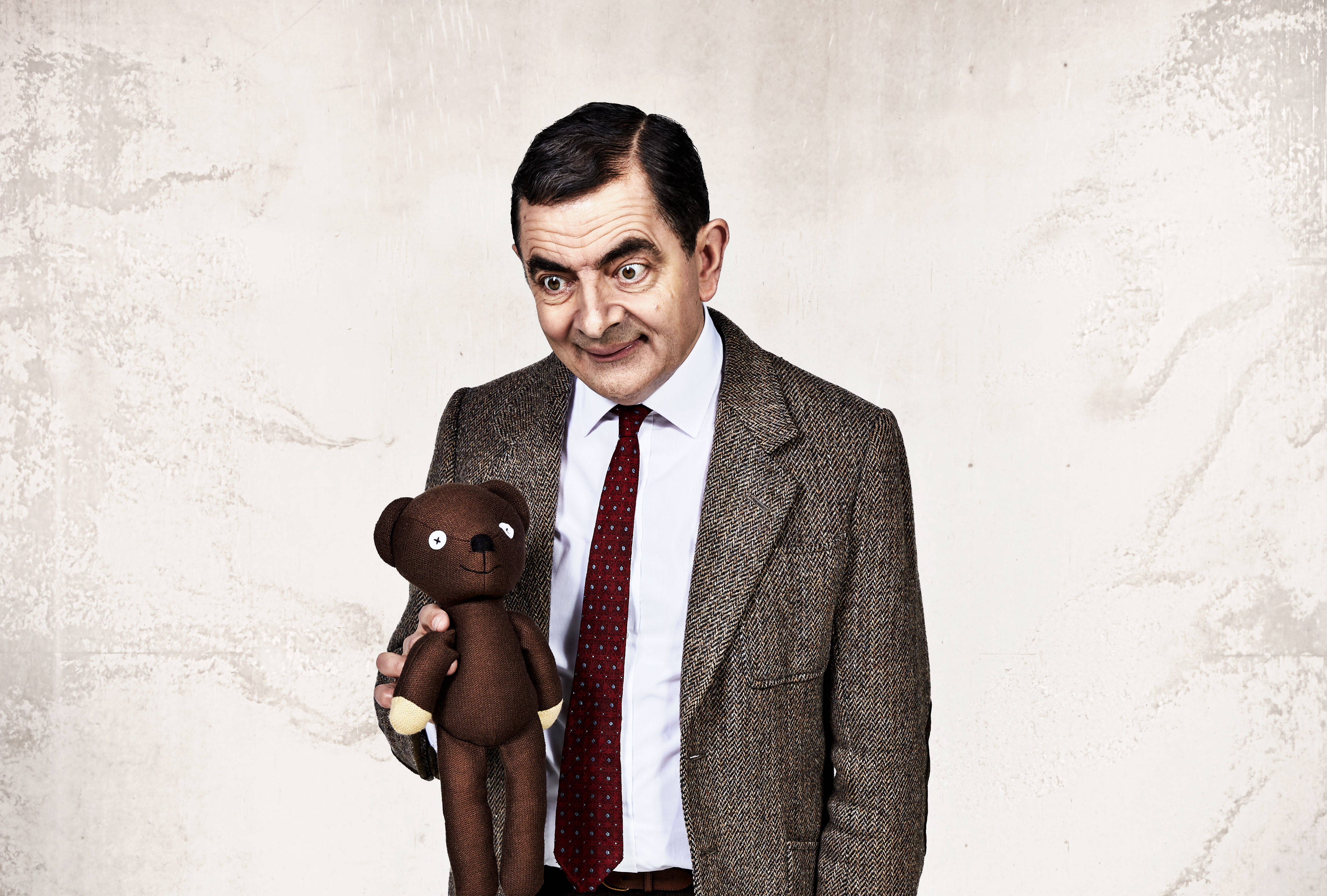 4K 桌布 壁紙 — Mr Bean 穿著粗花呢西裝，拿著一隻棕色泰迪熊，靠著有紋理的米白色牆壁，露出頑皮的笑容。
