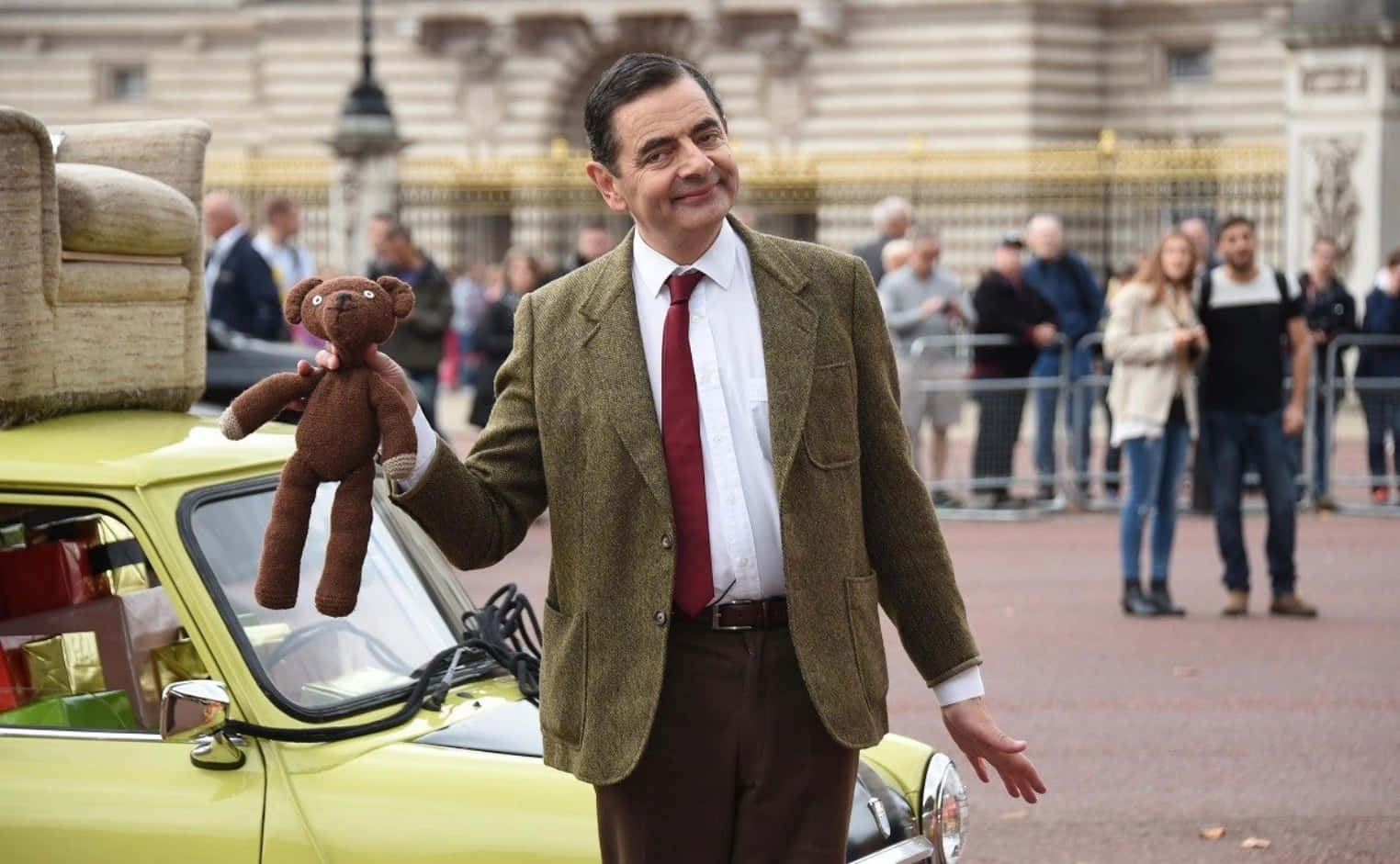 Mr Bean 桌布 壁紙 — Mr Bean 站在一輛黃色汽車旁邊，手持一隻泰迪熊，背後有一座宏偉的建築物。