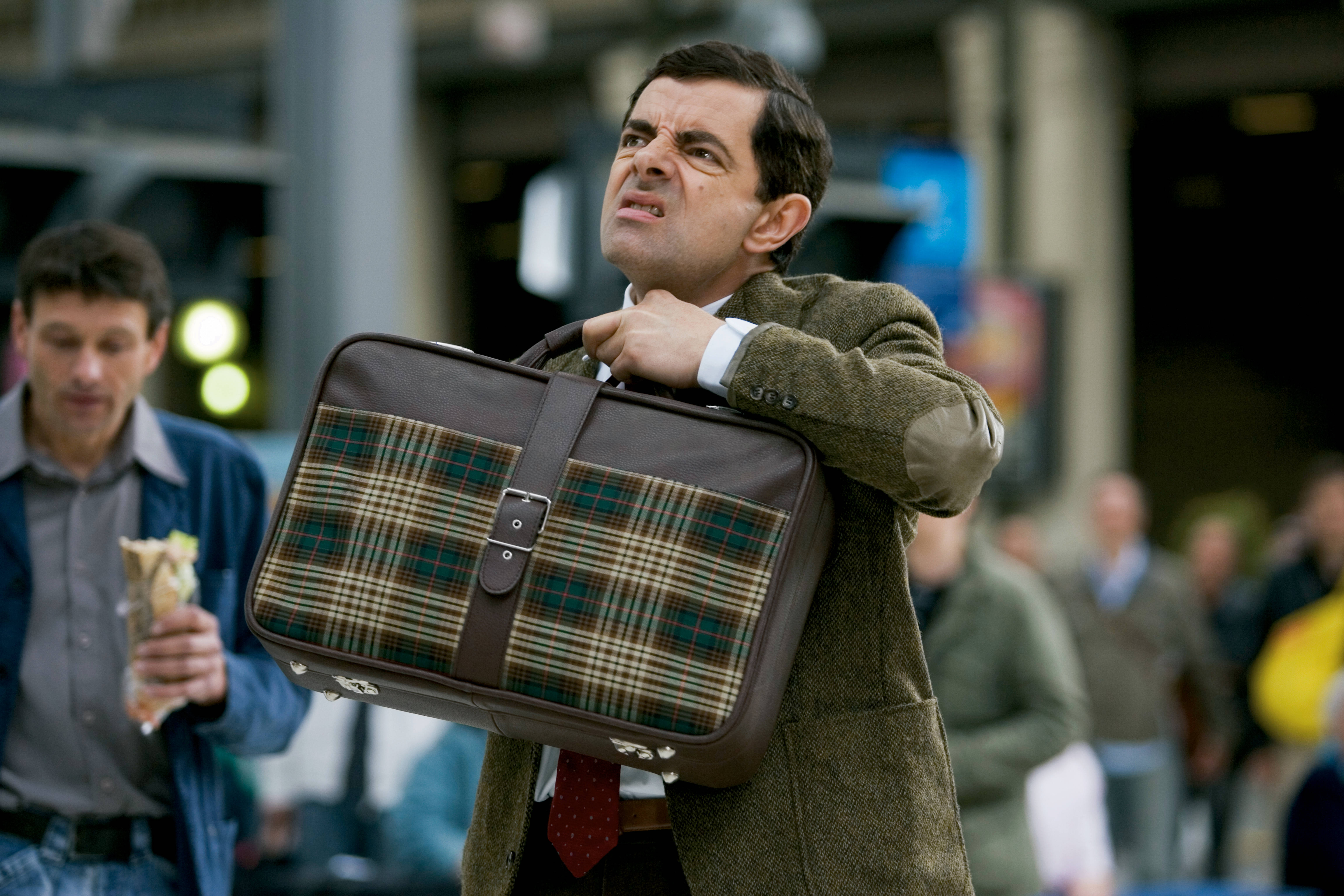 4K 桌布 壁紙 — Mr Bean 費力地拖著一個格仔行李箱穿過繁忙的城市車站，表情滑稽地繃緊。