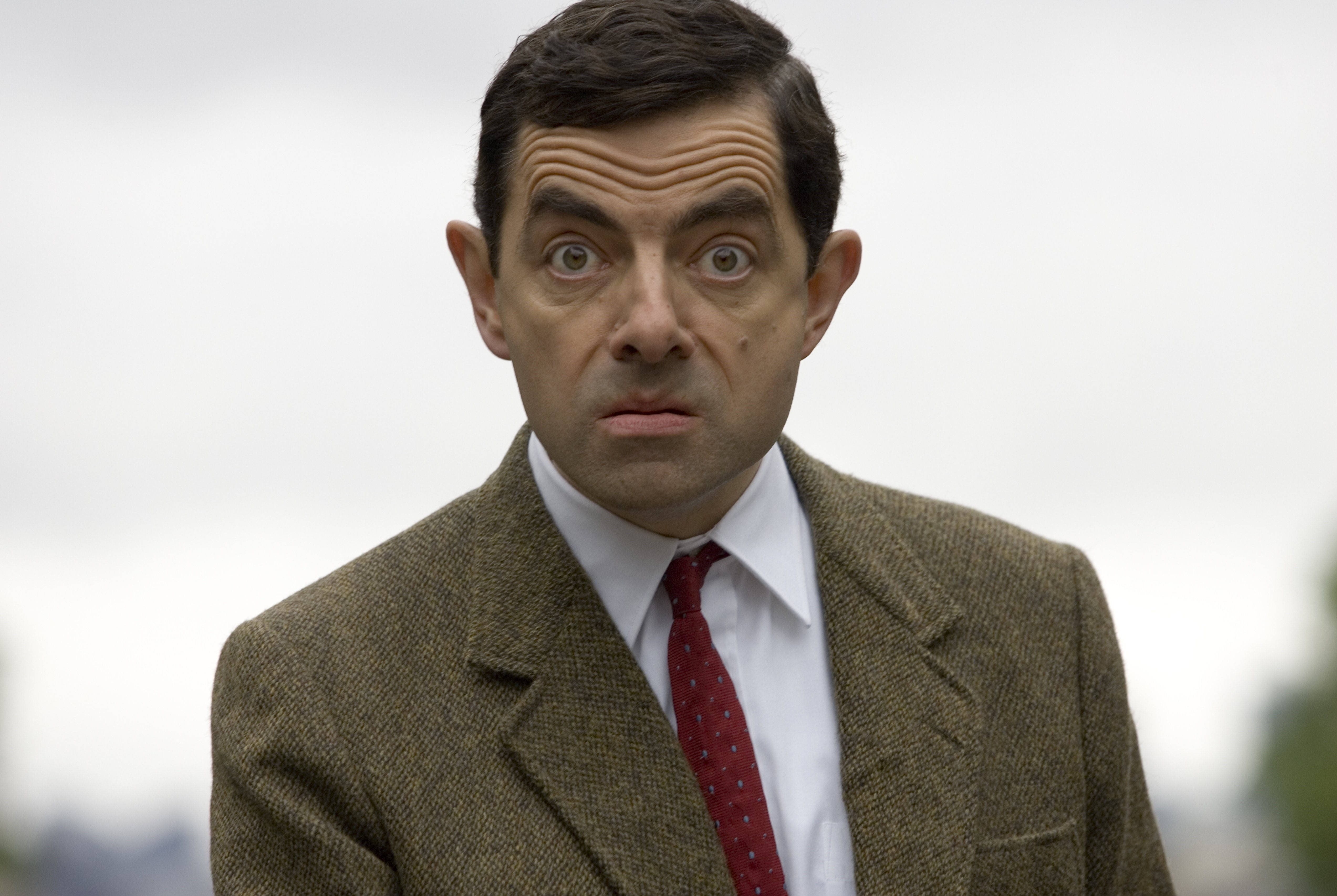 4K 桌布 壁紙 — Mr Bean 身穿棕色粗花呢西裝和紅色領帶，在模糊的戶外背景下瞪大眼睛震驚地凝視。