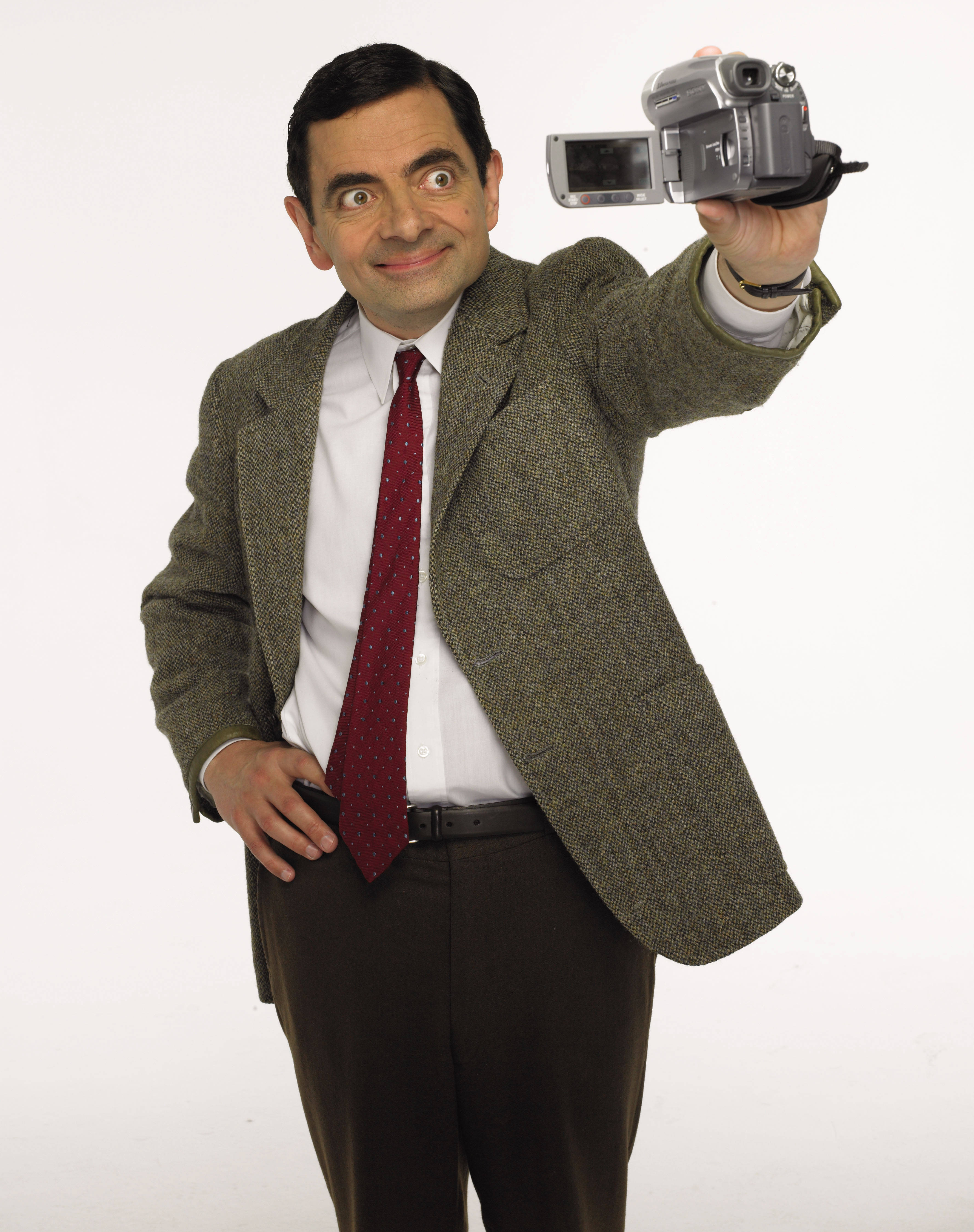  壁紙 — Mr. Bean 身穿粗花呢外套和紅色領帶，手持攝錄機，睜大眼睛露出頑皮的笑容，背景為純白色。