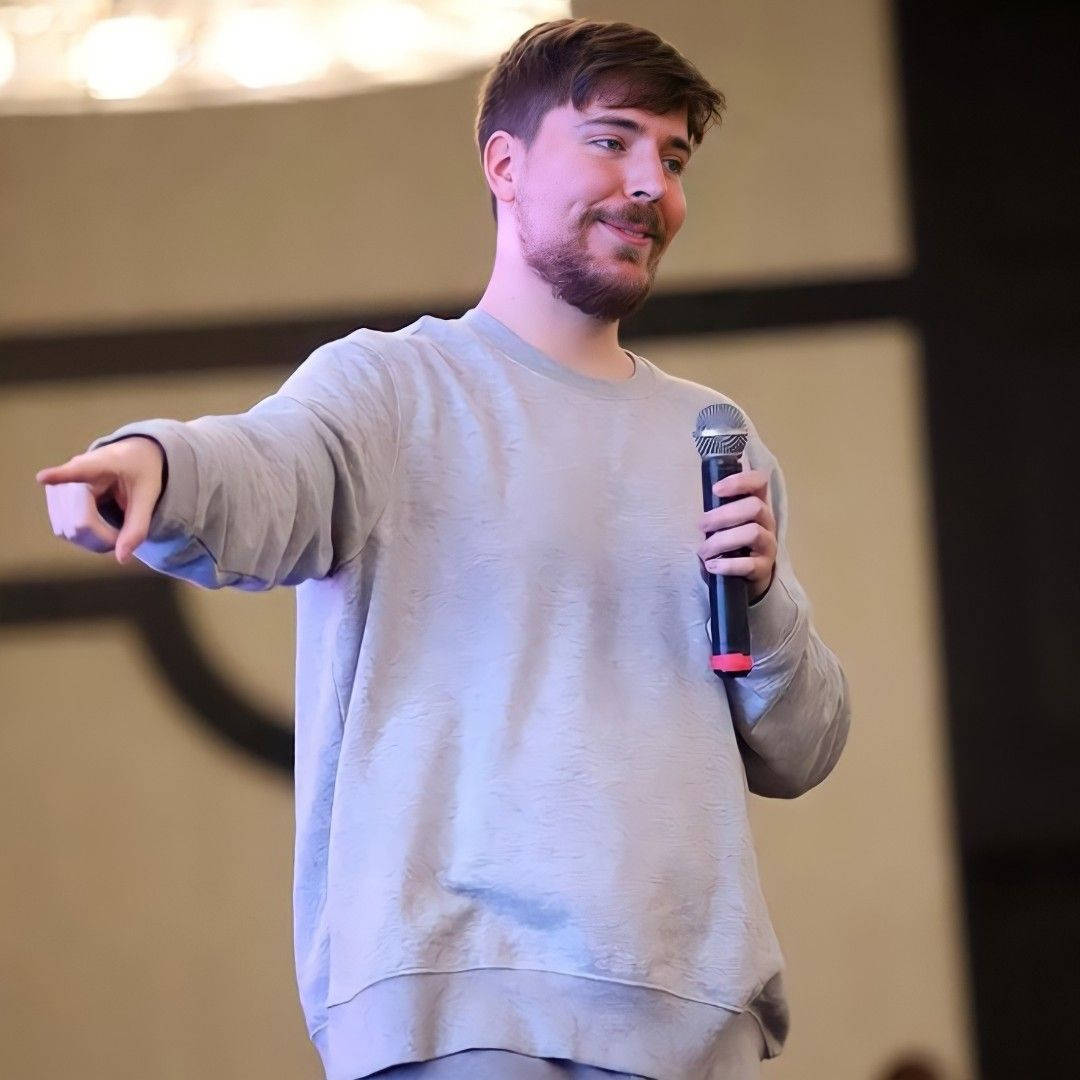 Mr. Beast 拿麥克風指向前方 壁紙 — 一位留鬍鬚的微笑男子拿著咪高峰，在溫暖燈光的舞台上邊說話邊做手勢。