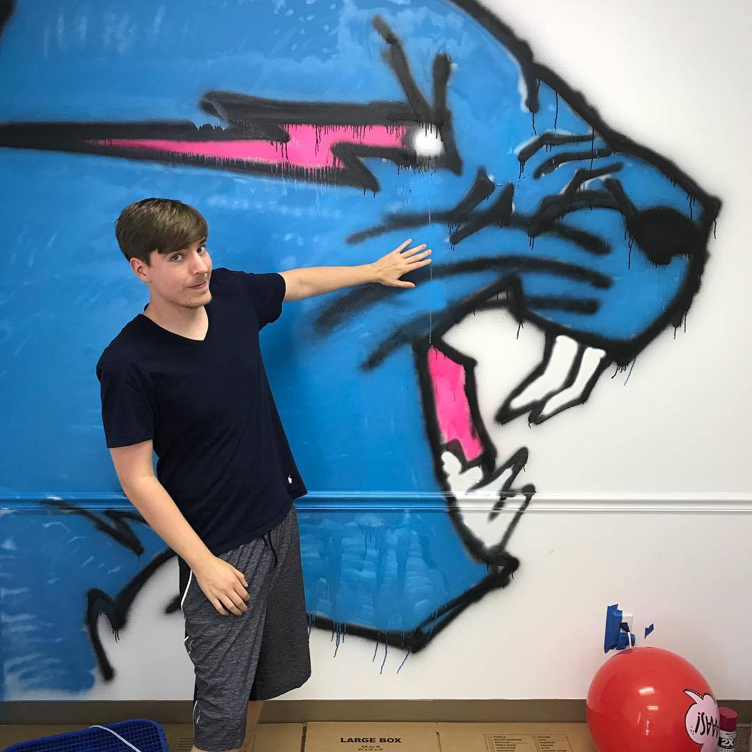 Mr Beast 噴漆標誌 壁紙 — 一名年輕男子站在白色牆壁上，旁邊是充滿活力的藍色和粉紅色塗鴉壁畫，畫著一隻咆哮的動物頭部。