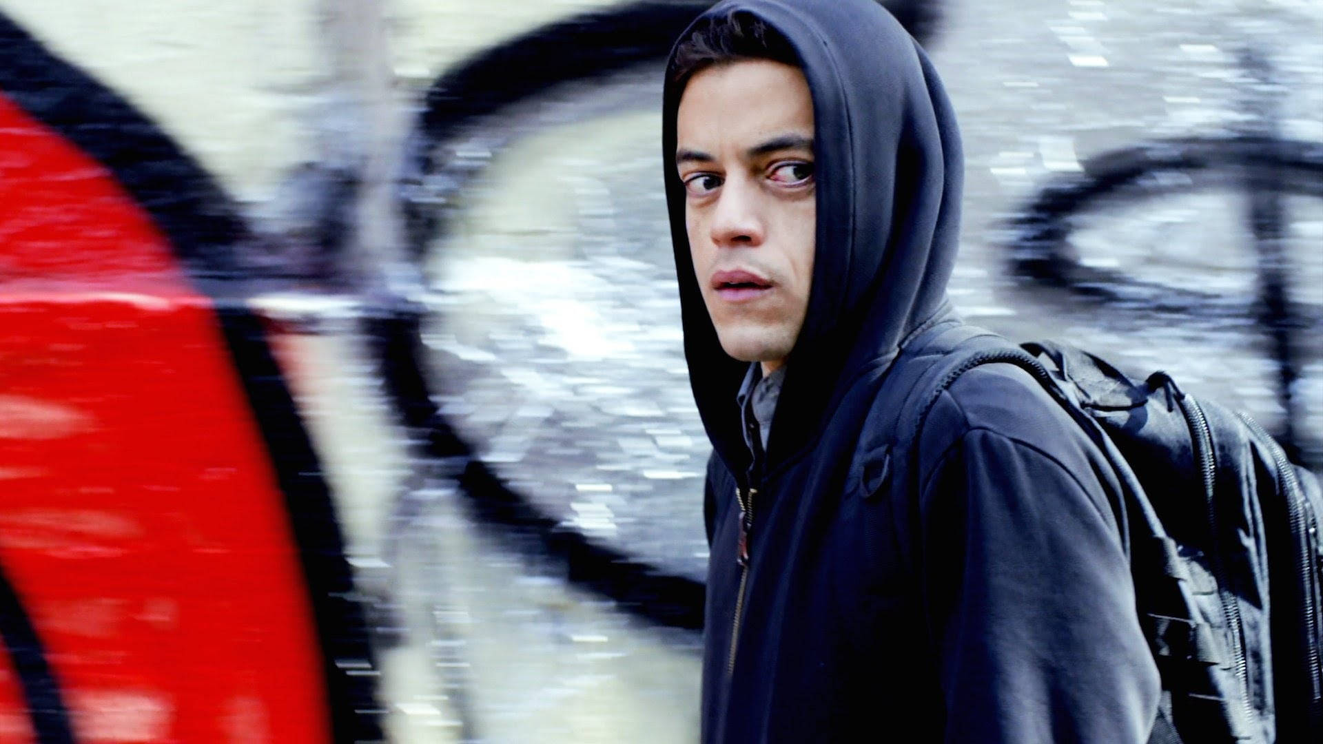 Mr. Robot 下一季預告 壁紙 — 一個穿著黑色連帽衫和背包的年輕男子，凝視著一幅模糊的塗鴉牆，上面有醒目的紅色和黑色標籤。