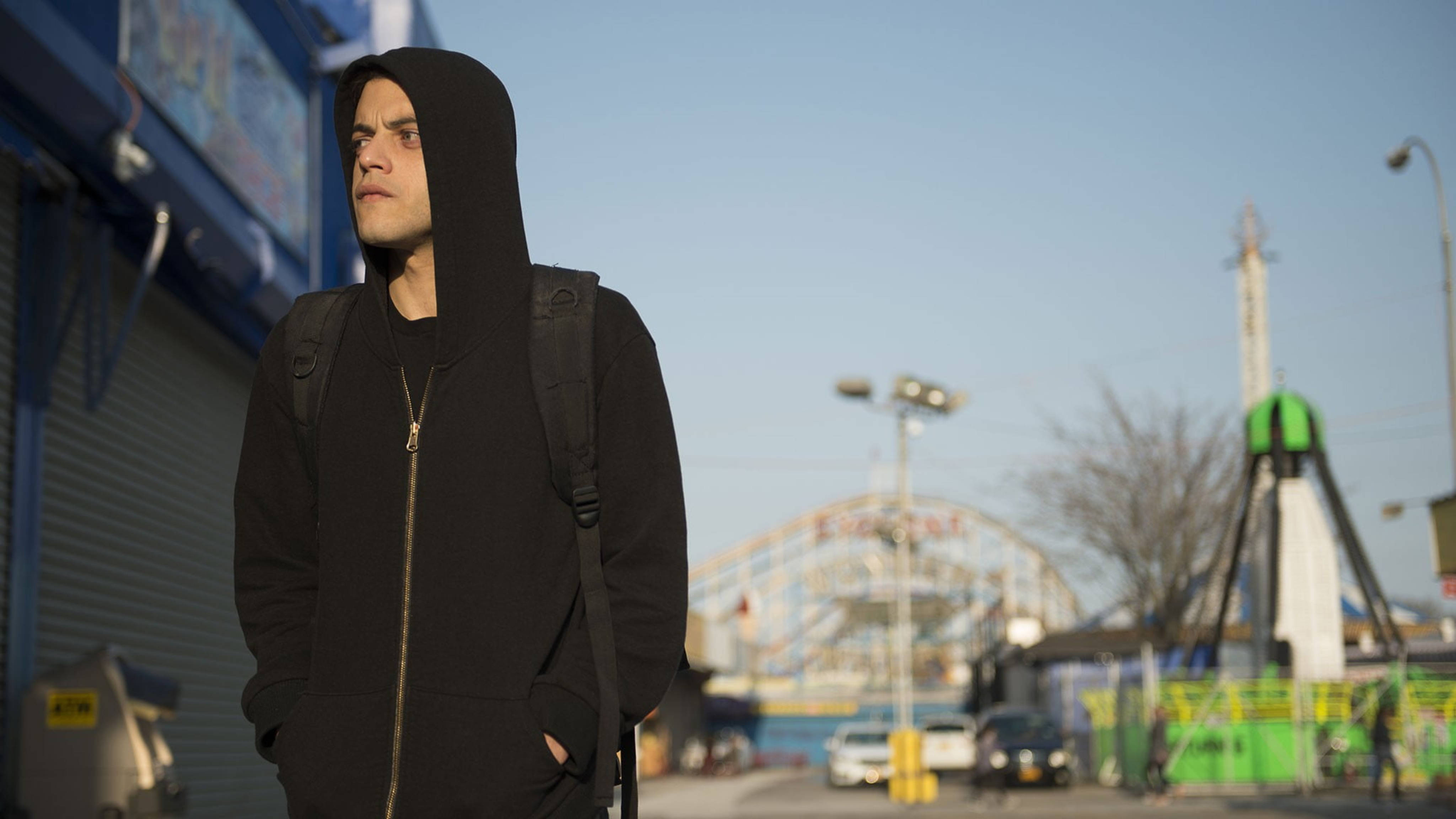 Mr. Robot 在城市街道 壁紙 — 一個穿著黑色連帽衫的年輕男子在陽光明媚的城市街道上沉思，背景有嘉年華。