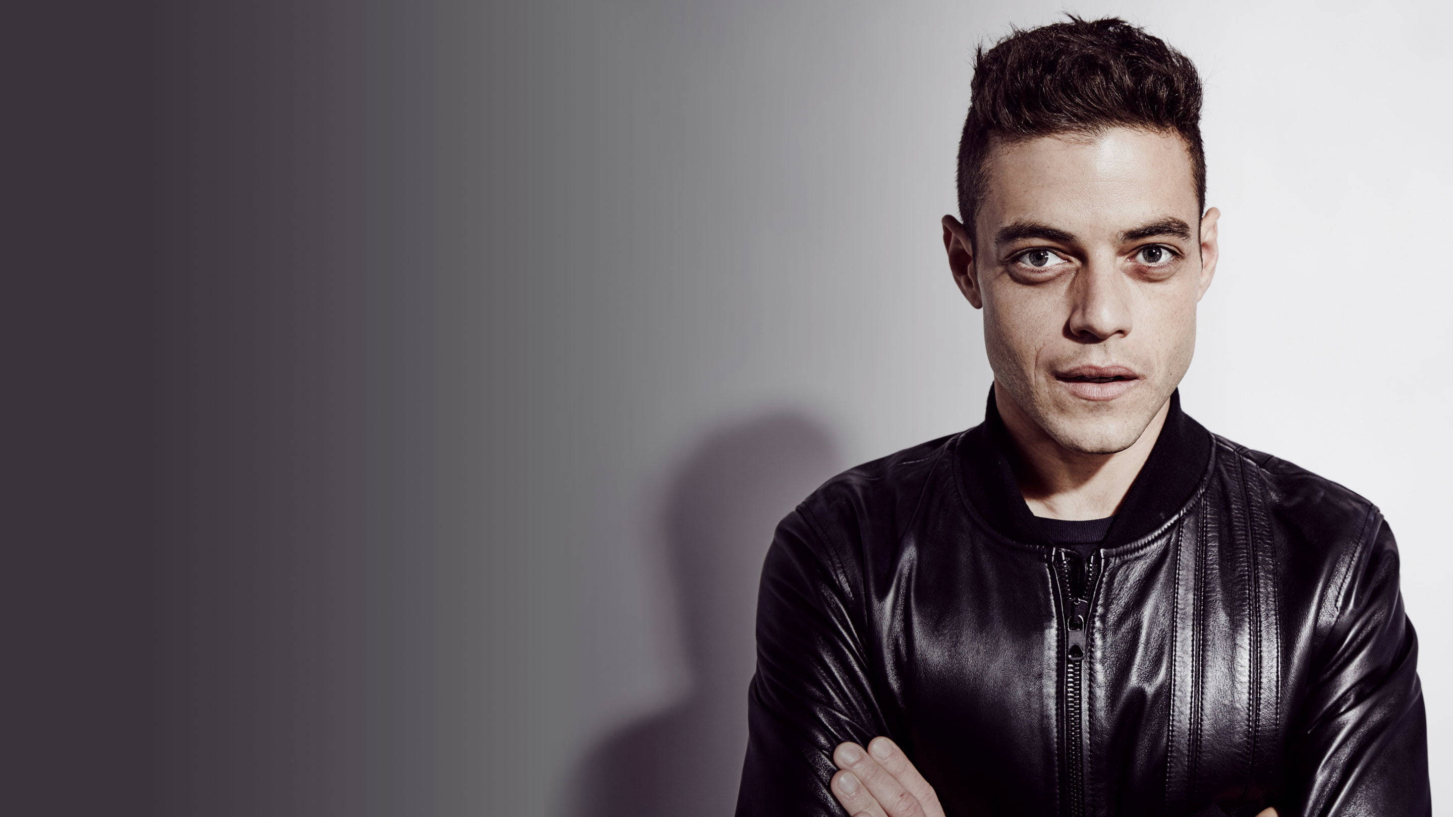 《Mr. Robot》演員拉米·馬利克 壁紙 — 一個穿著黑色皮褸的男人自信地交叉雙臂，站在簡約灰色漸變背景前。