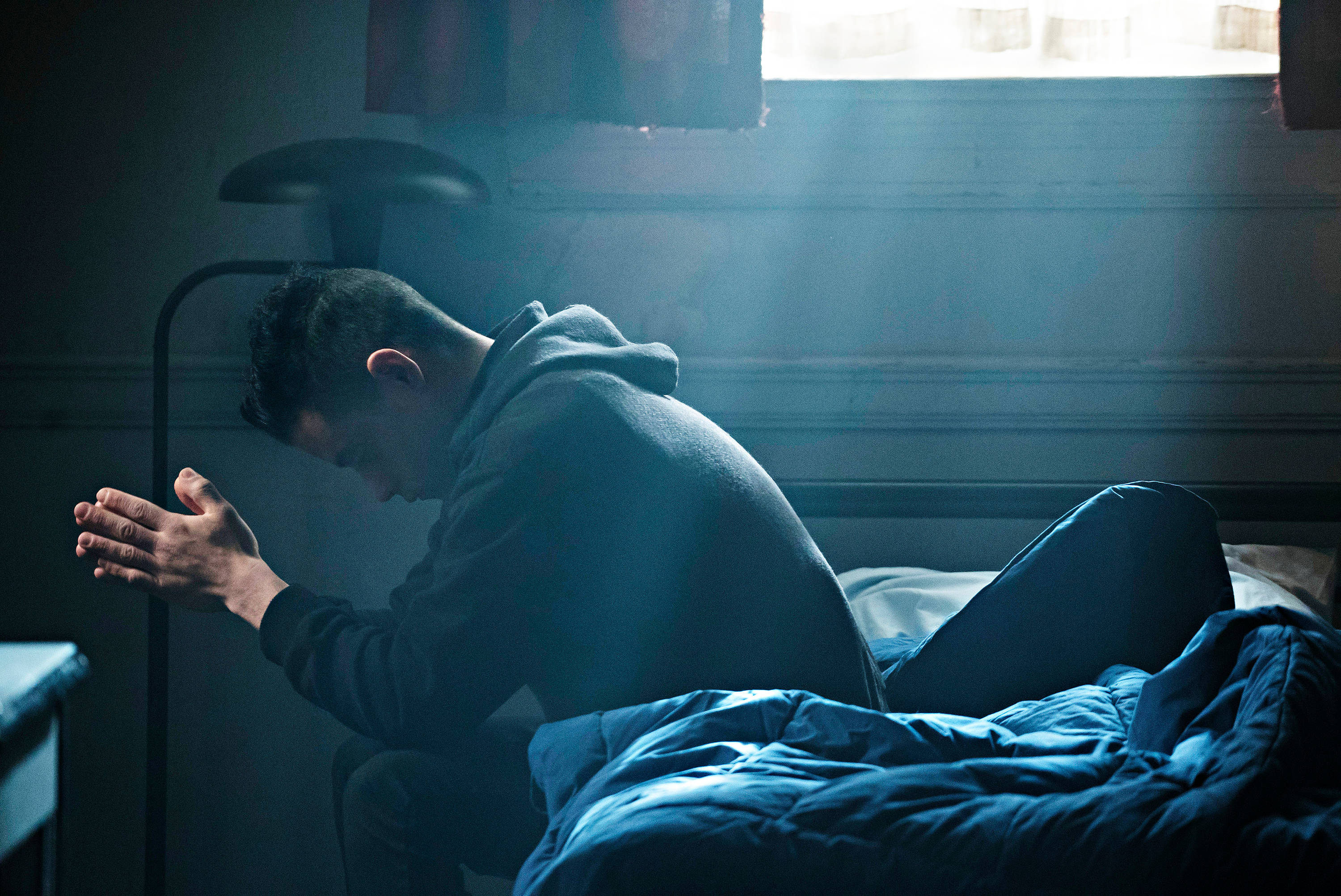 《Mr. Robot》演員拉米·馬勒克 壁紙 — 一個男人跪在床邊，房間昏暗，陽光透過窗戶照射進來，投下戲劇性的陰影。