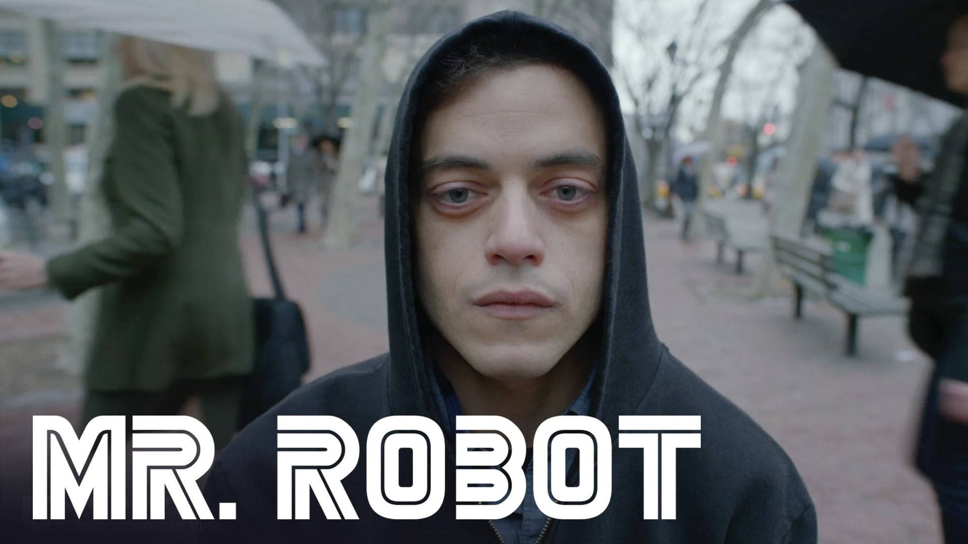 《Mr. Robot》艾略特·第二季 壁紙 — 一個穿著黑色連帽衫的男人在雨天的城市人行道上，背景是模糊的行人和光禿禿的樹木，他正強烈地凝視著鏡頭。