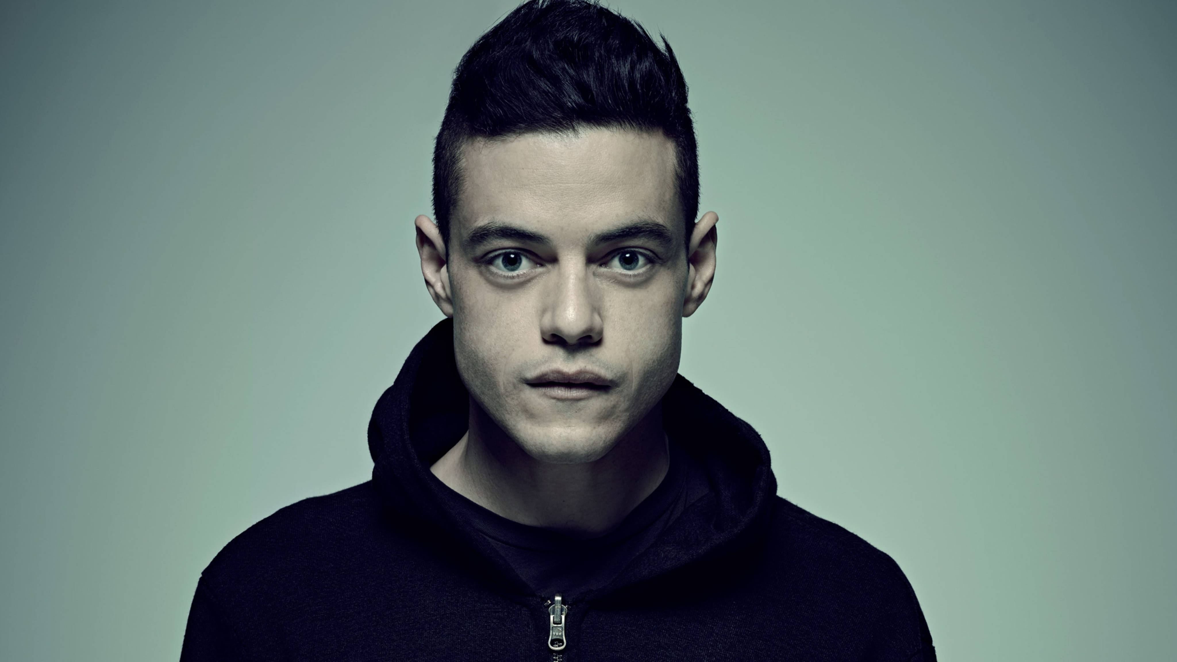 《Mr. Robot》艾略特·阿爾德森 壁紙 — 一個眼神銳利的年輕男子直接凝視鏡頭，身穿黑色連帽衫，背景為柔和的灰色。