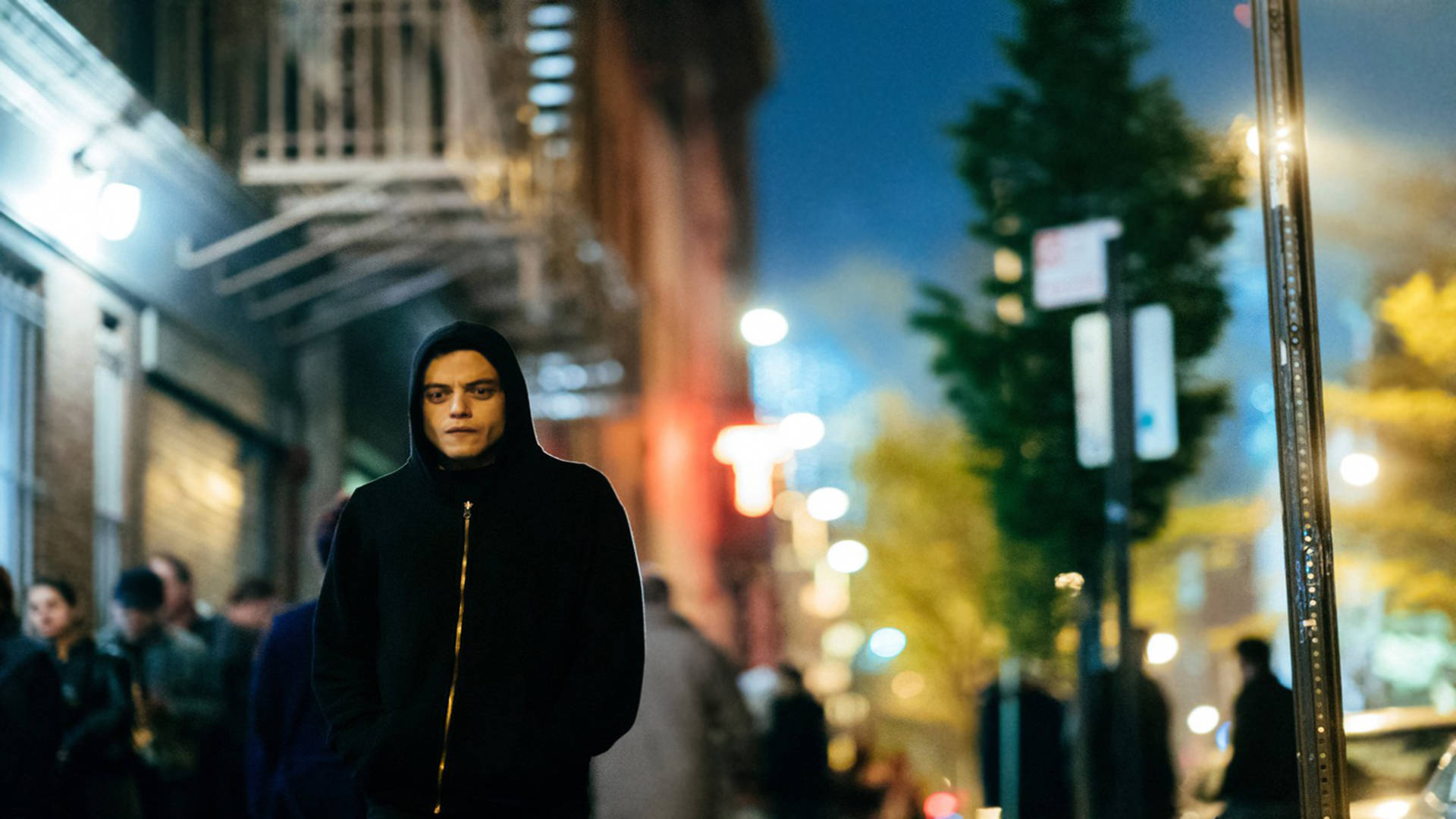 《Mr. Robot》艾略特夜間街頭 壁紙 — 一個穿著黑色連帽衫的年輕男子夜晚獨自站在繁忙的城市街道上，被溫暖的路燈和發光的招牌照亮。