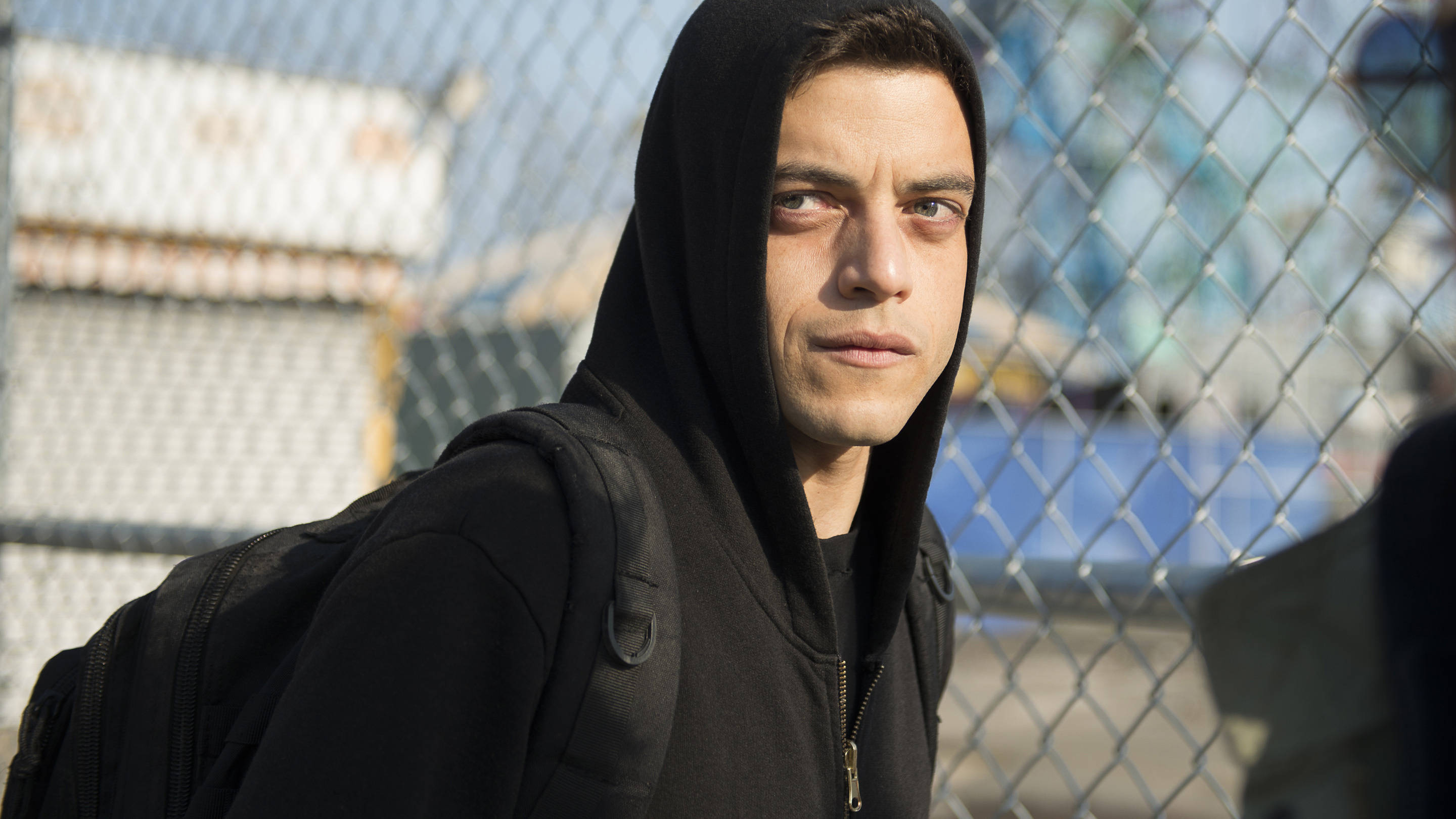 《Mr. Robot》艾略特靠近鋼鐵柵欄 壁紙 — 一個穿著黑色連帽衫和背包的年輕男子在明亮的日光下透過鐵絲網凝視。