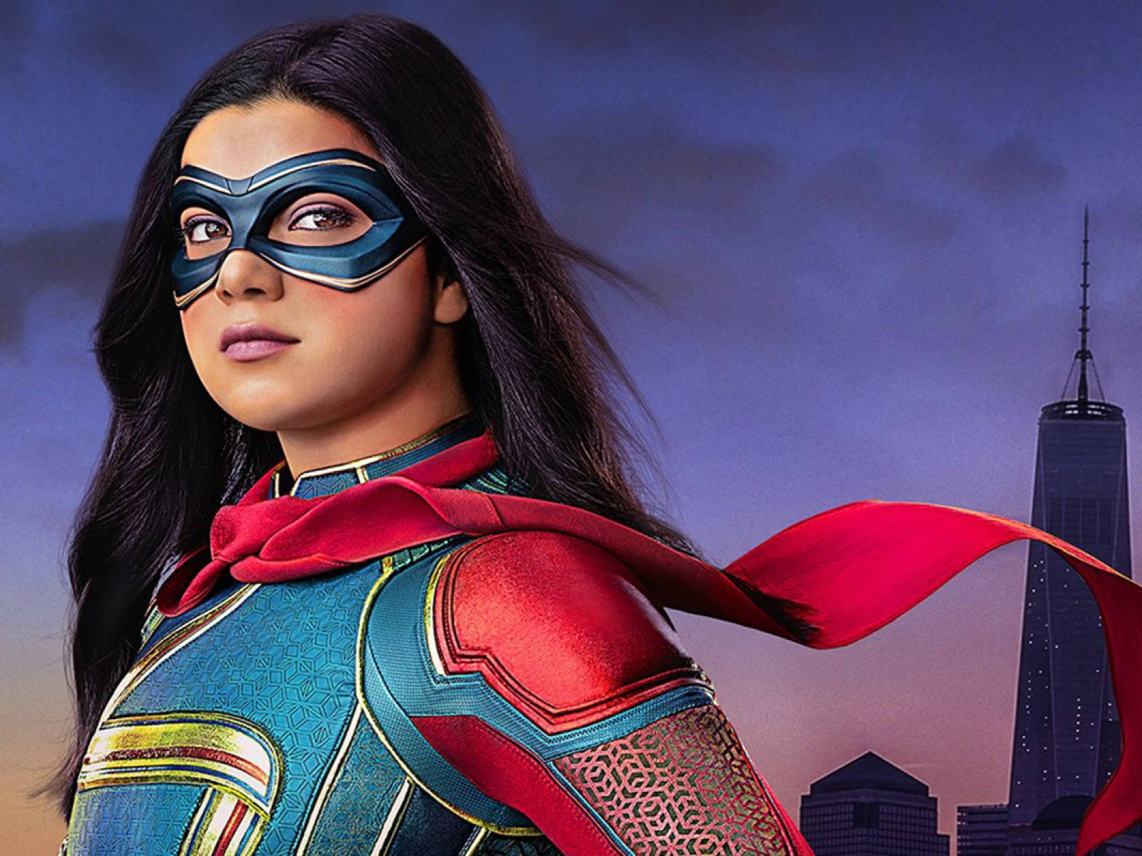 Ms Marvel 伊曼·維拉尼 壁紙 — 一位身穿藍紅緊身衣、披著飄逸斗篷的女超級英雄，在黃昏的城市天際線前自信地站立。