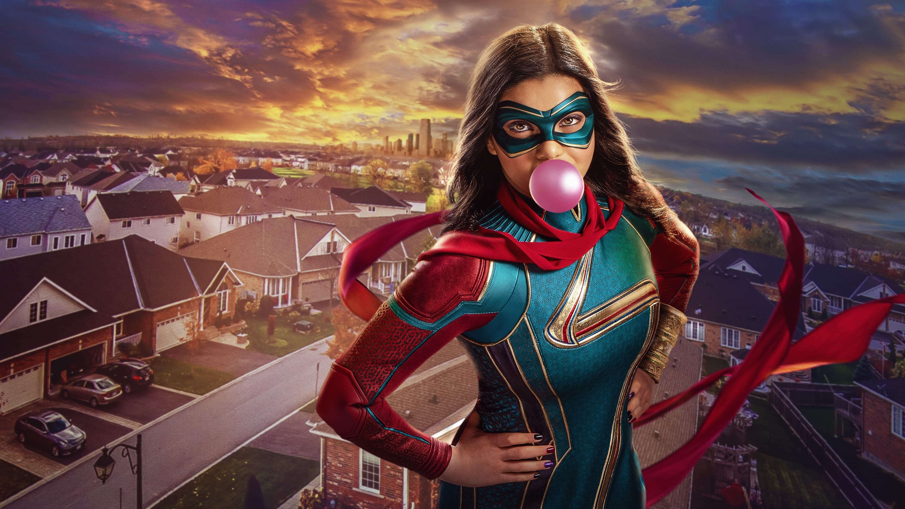 Ms Marvel 動作中的鼓舞力量 壁紙 — 一位身穿青綠色和紅色服裝的女超級英雄在日落時分在郊區上空吹出粉紅色泡泡糖泡泡。