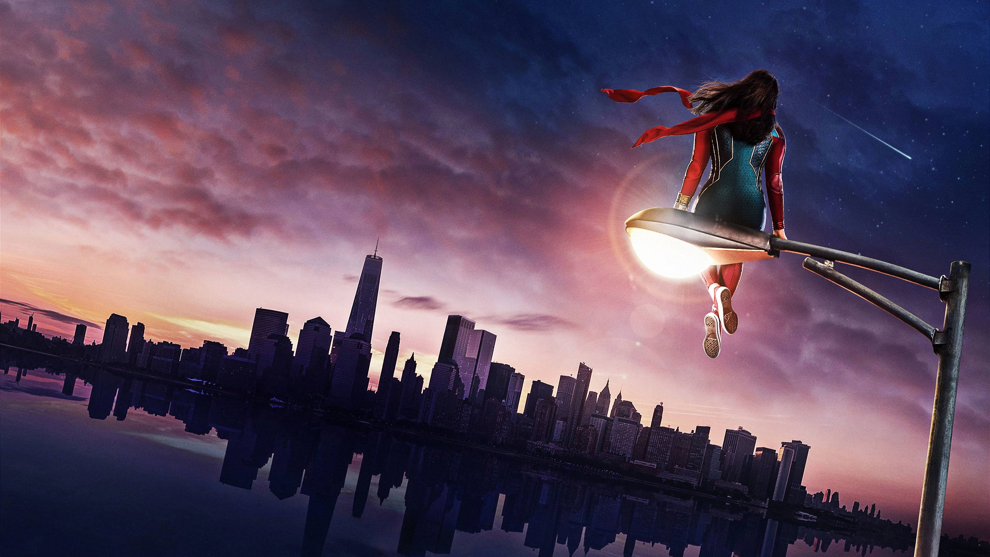 Ms Marvel 坐在街燈柱上 壁紙 — 一位身穿紅藍服裝的女超級英雄從發光的路燈上躍下，背景是水中倒映的充滿活力的暮色城市景觀。