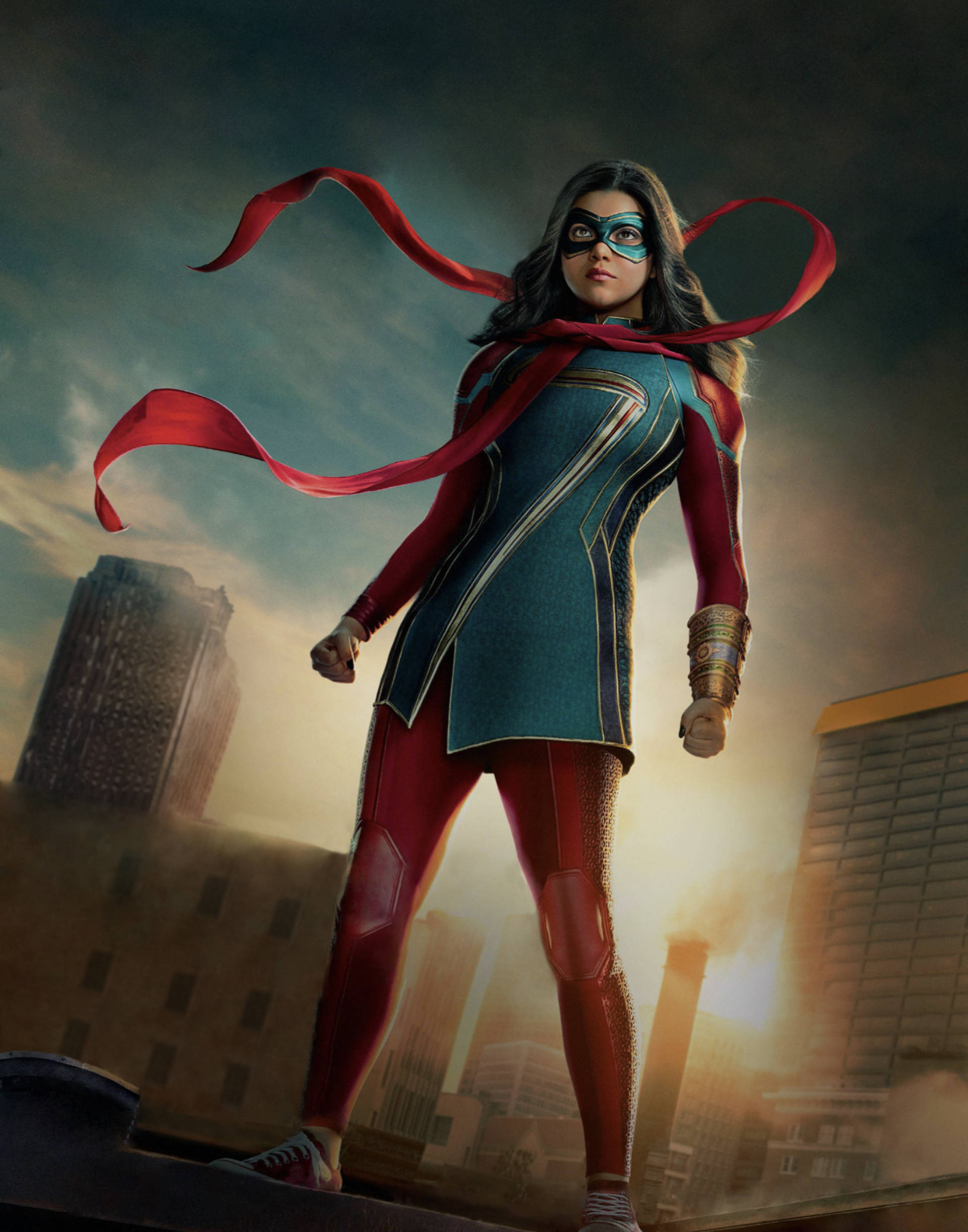 Ms Marvel 日落時分 壁紙 — 一位身穿紅藍服裝的女超級英雄自信地站在城市建築物頂端，披風在風中飄揚，背景是戲劇性的天空。