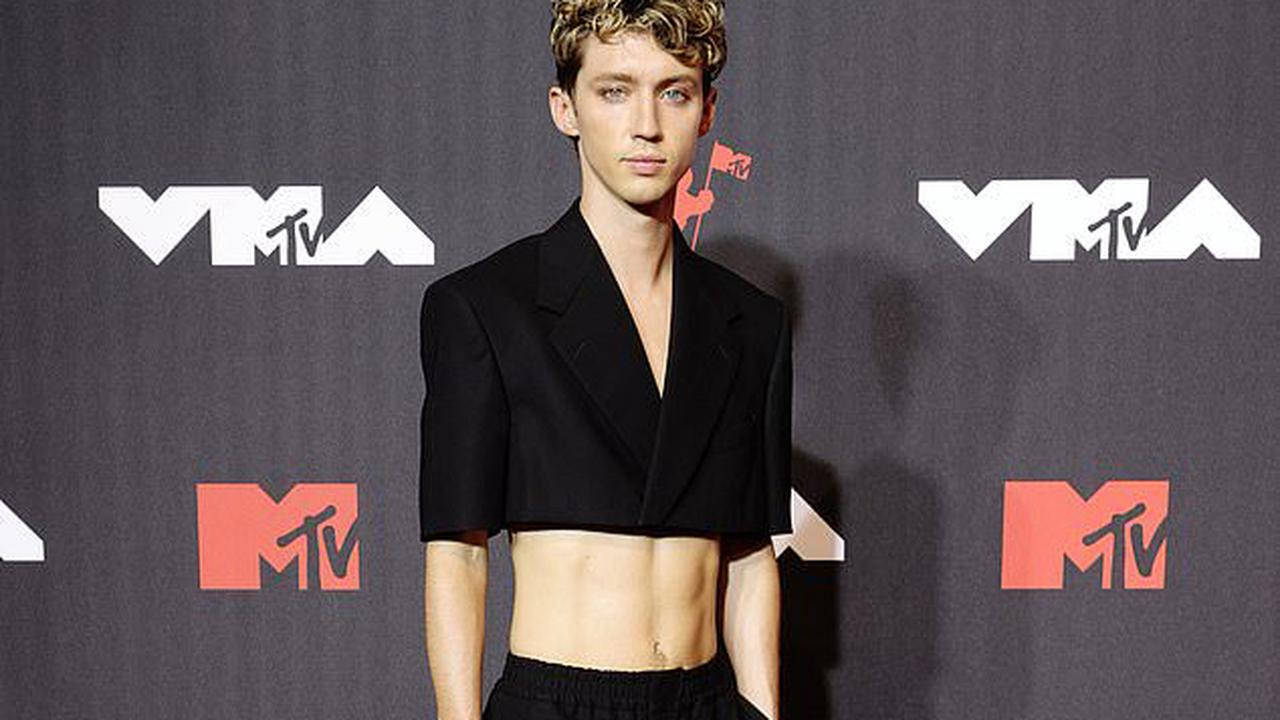 特洛伊·西文出席MTV音樂錄影帶大獎 壁紙 — 一位捲髮年輕男子穿著黑色短版西裝外套和黑色褲子，背景是深灰色的MTV VMA背景。