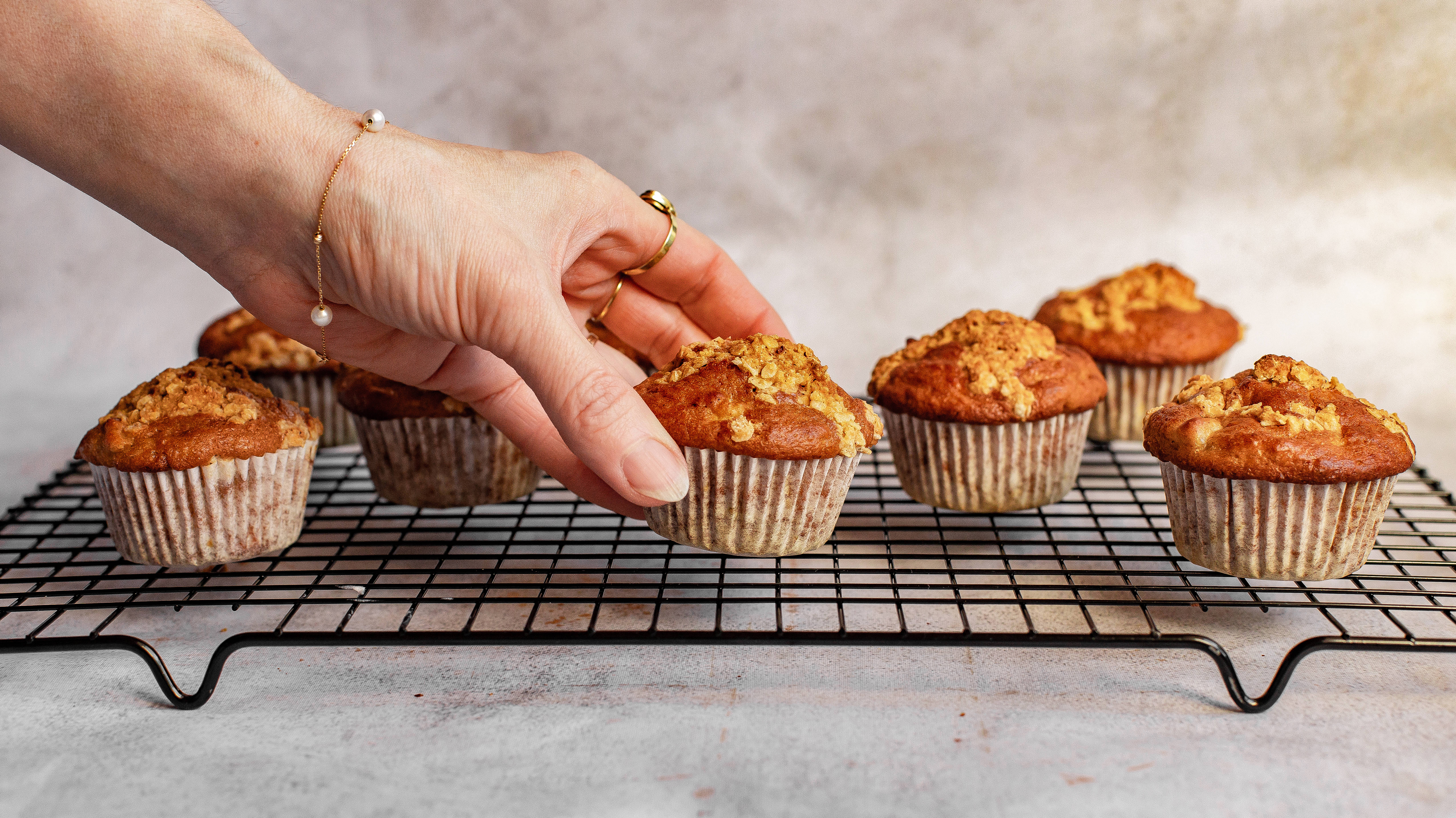 杏桃烤色 muffin 壁紙 — 一隻戴著金戒指的手輕輕地從黑色金屬冷卻架上拿起一個溫暖的碎屑頂鬆餅。